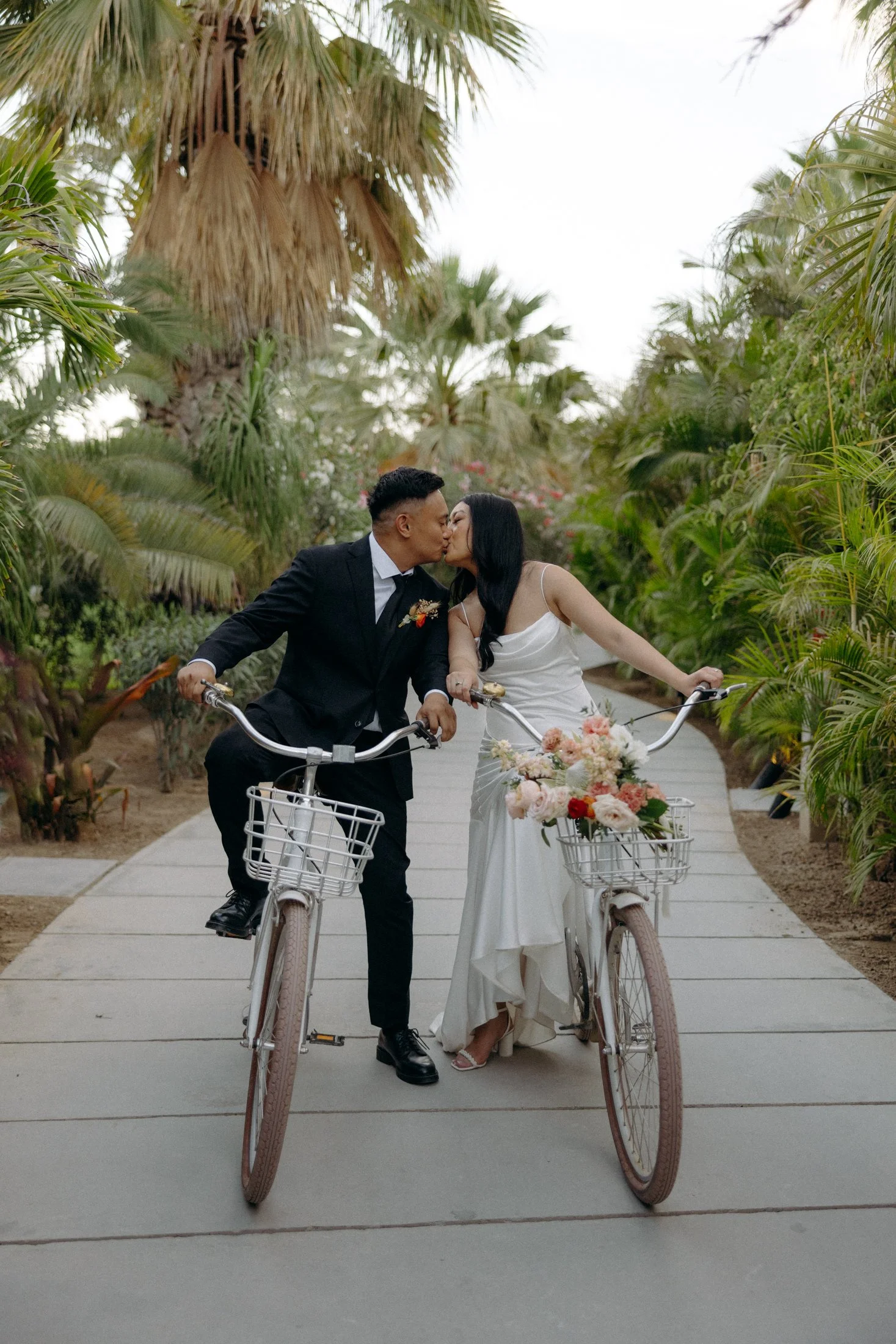 a wedding elopement couple kisses on their bikes on a path through a forest of palm trees at the acre resort in cabo, mexico