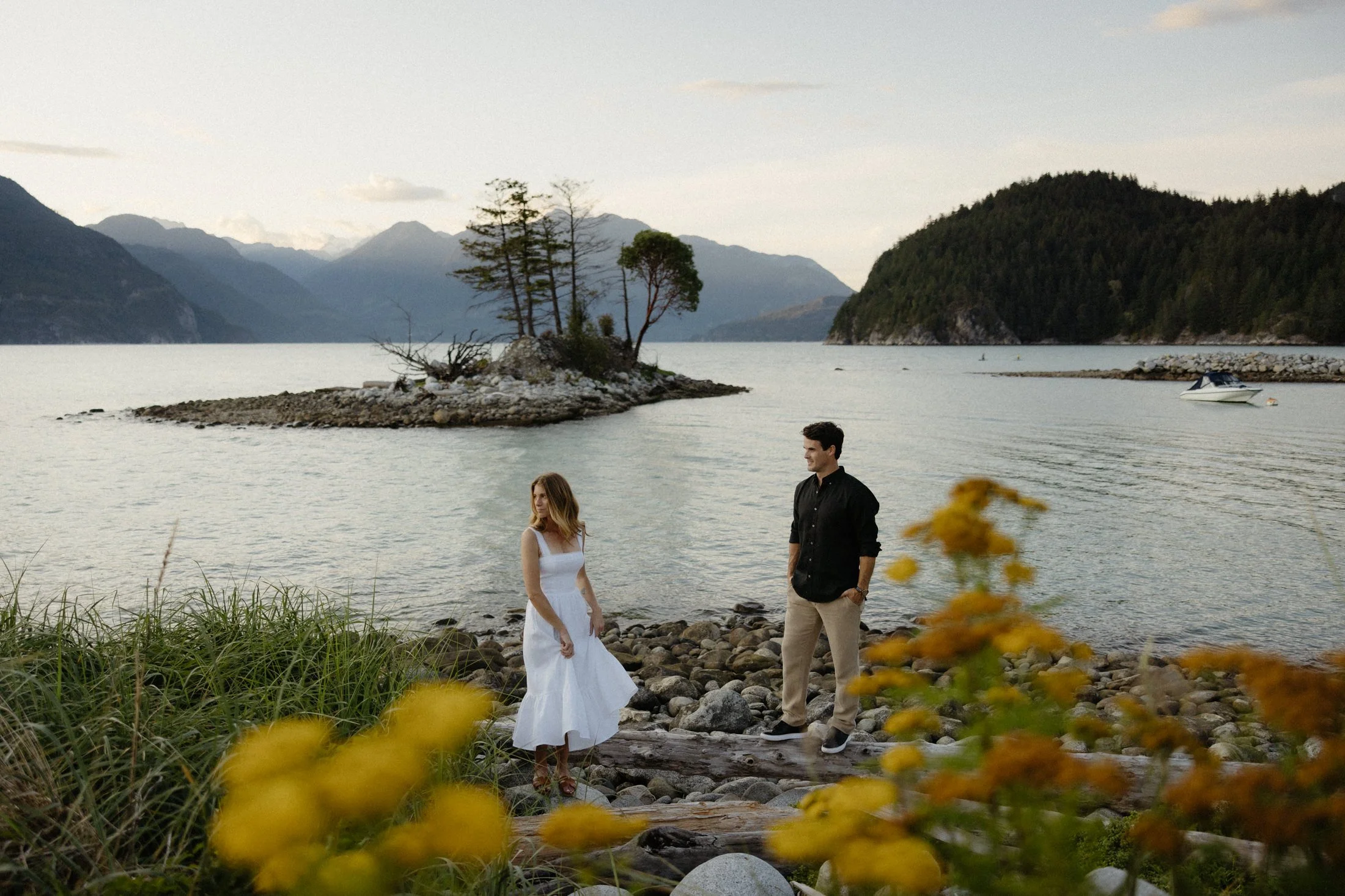 A couple stands separately looking to the left, through some yellow flowers with a small rocky island in the background complete with the ocean and mountains