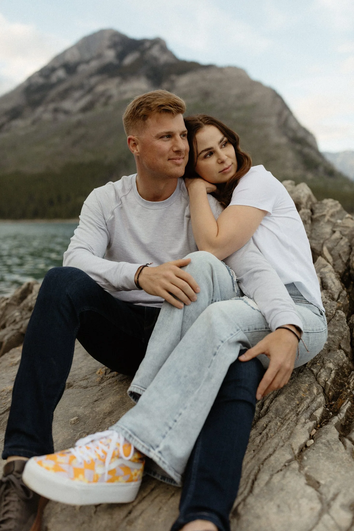 a couple cuddles up on a rock and looks into the distance with a lake and mountain in the background in banff national park