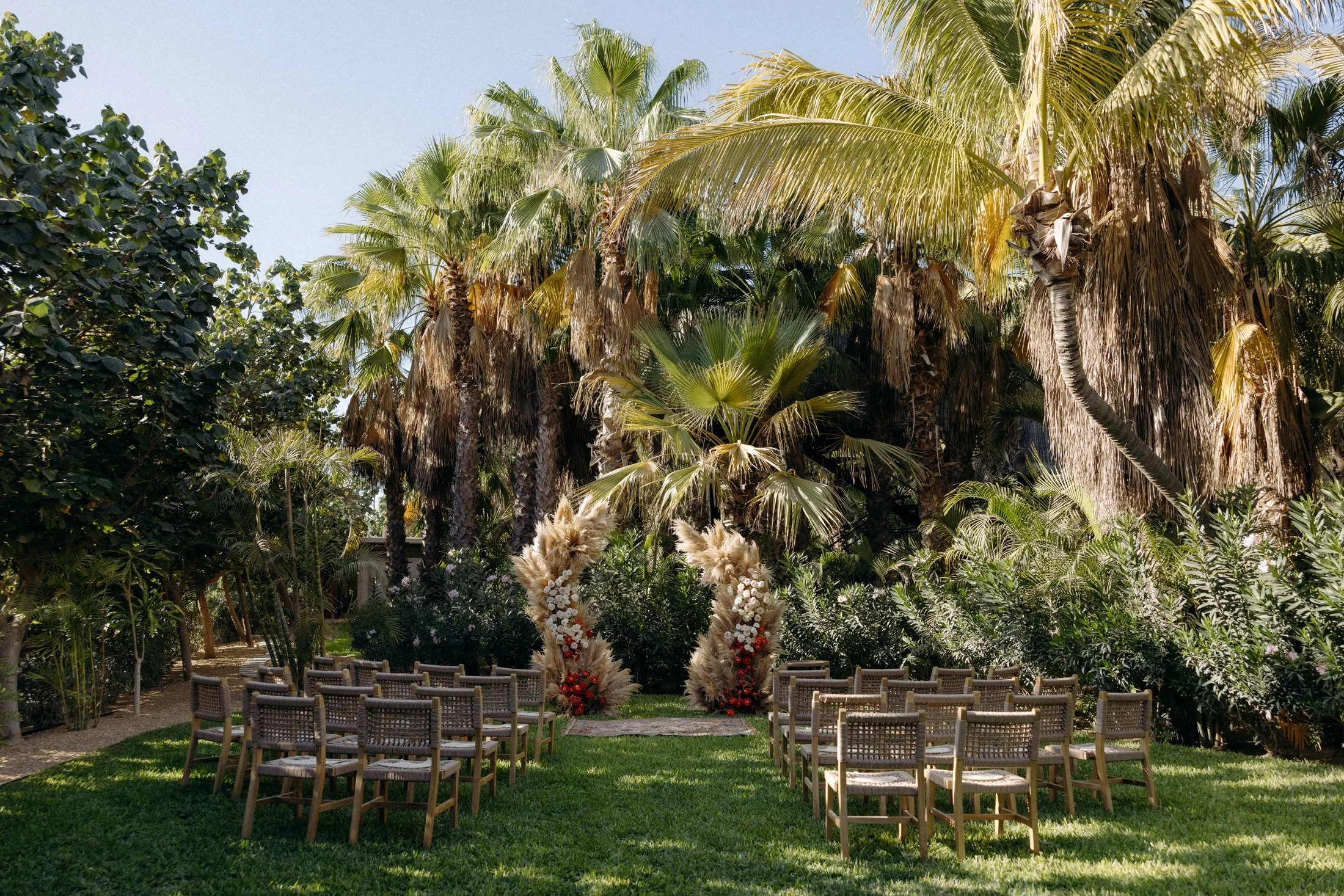 A view of the wedding ceremony space on lover's lawn with green grass, natural-looking chairs, and a floral arch in front of a forest of palm trees at acre resort in Cabo, Mexico