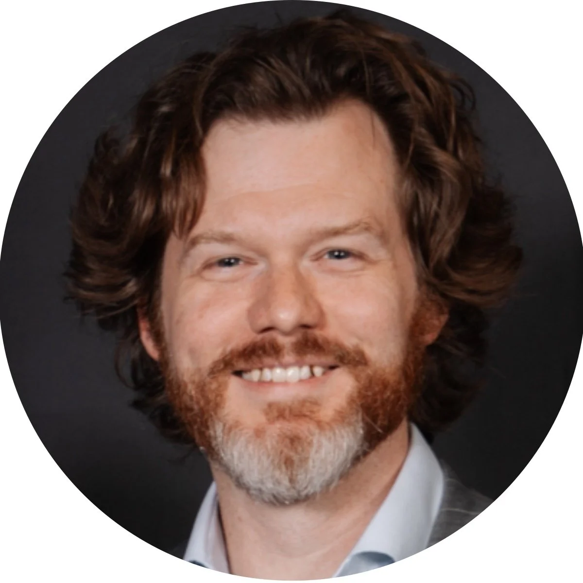 Close-up photo of a smiling man with medium-length curly brown hair, a beard, and blue eyes, wearing a suit jacket and white shirt against a dark background.