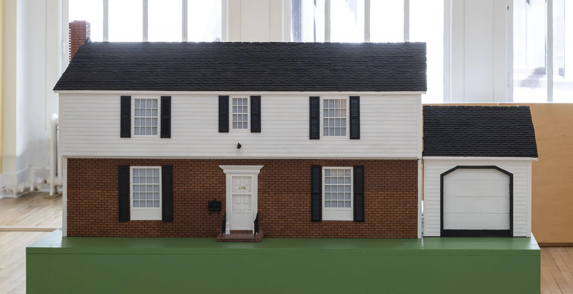  JONA FRANK Model Home, 2021 Custom replica of a suburban house. Plywood, Mini-roof shingle, paint, wire, Luma mini projector. Showing a slideshow, 14:54 minutes of Jona Frank’s family photographs and footage of the Exploding House. 33 x 62 x 28 in  