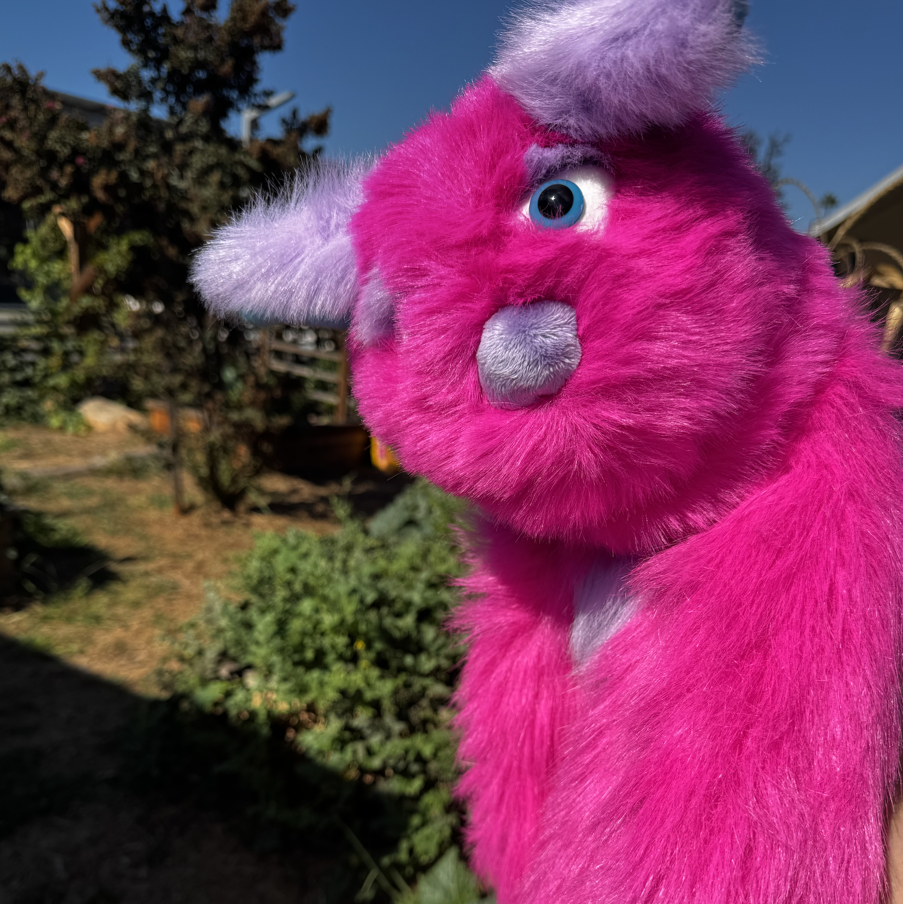 Close-up of a pink furry puppet with purple ears, blue eyes, a purple nose, and a light purple chin, outdoors in a garden area with trees and plants in the background under a clear blue sky.