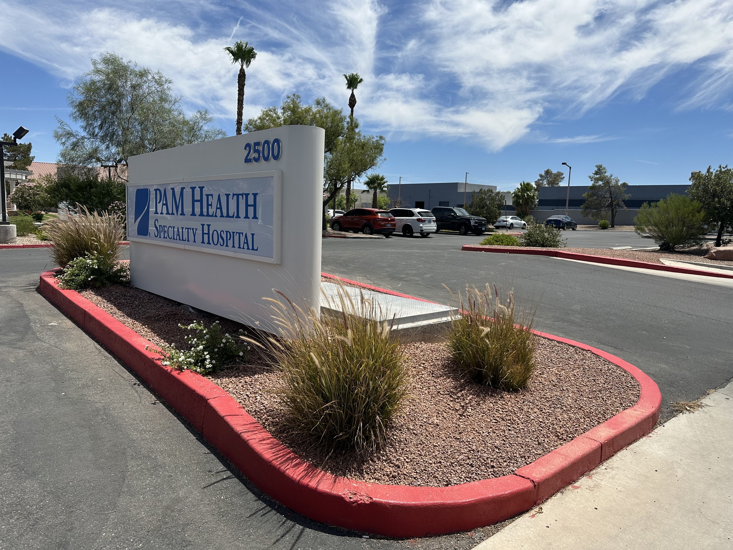 Sign for Pam Health Specialty Hospital at 2500, with a parking lot and trees in the background on a sunny day.