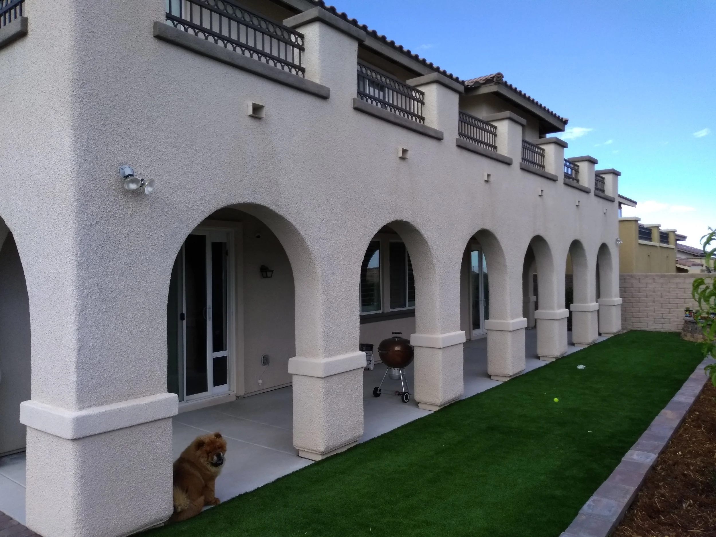 The back porch of a large beige stucco house with multiple rounded arches, two sliding glass doors, and a second-floor balcony with a metal railing. There is a small brown dog sitting on the patio near one of the arches, a charcoal grill, and two ten