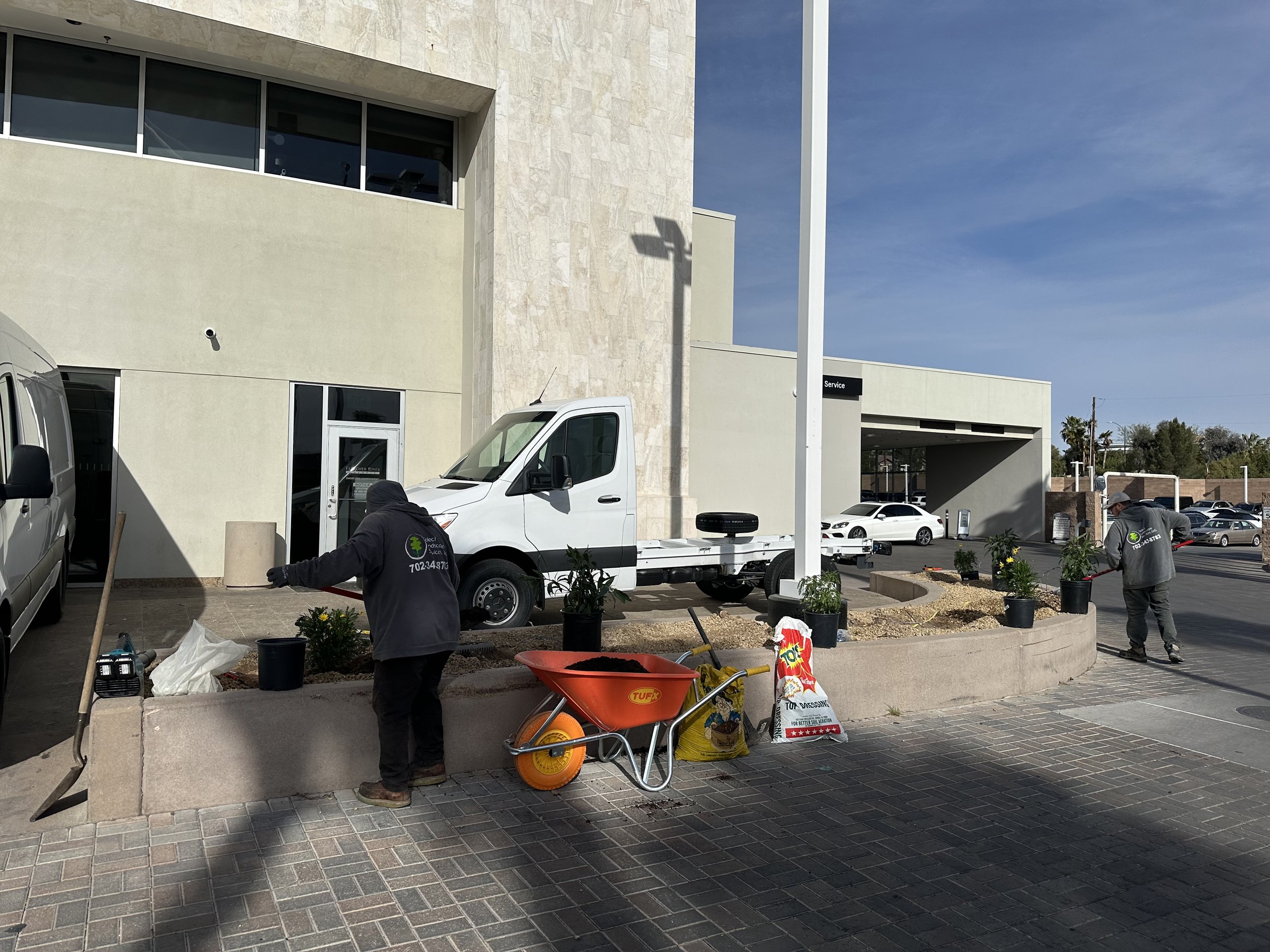 Two workers planting flowers in a small landscaped area at a commercial building with a white van parked nearby