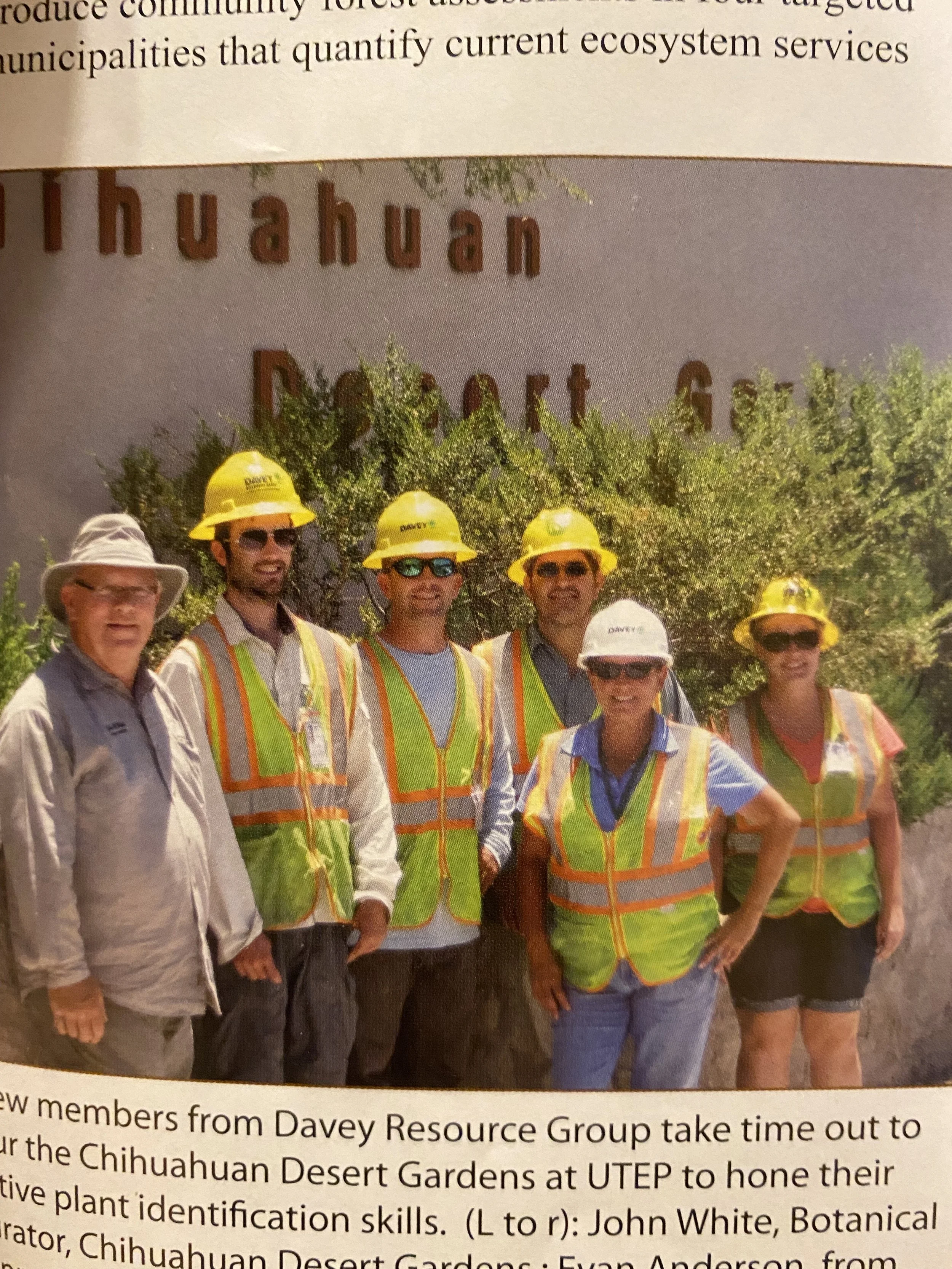 Group of six people wearing hard hats and safety vests standing outdoors in front of a sign that reads 'Chihuahua Desert Gardens'.