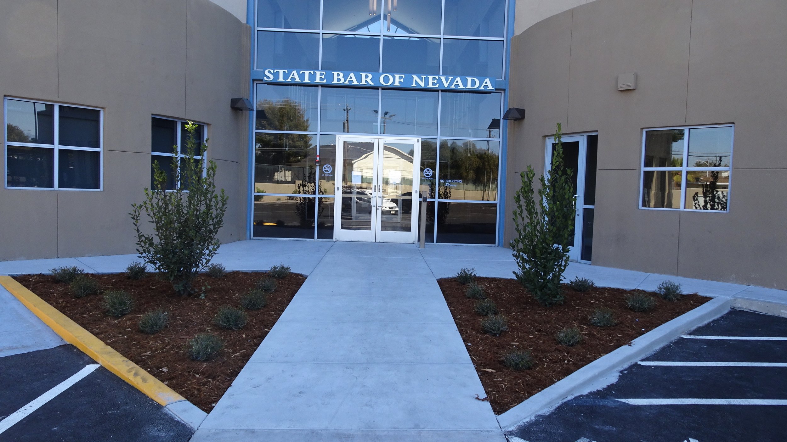 Exterior of a building with a sign that reads 'State Bar of Nevada' above glass doors. There are bushes and plants on either side of the walkway leading to the entrance, with a parking lot visible in the foreground.