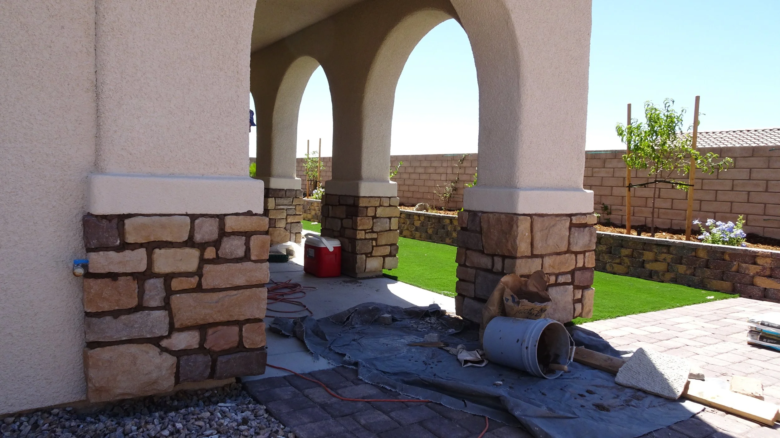Under-construction porch with stone columns and arches, outdoor gardening area with trees and brick wall, construction materials, and tools on ground.
