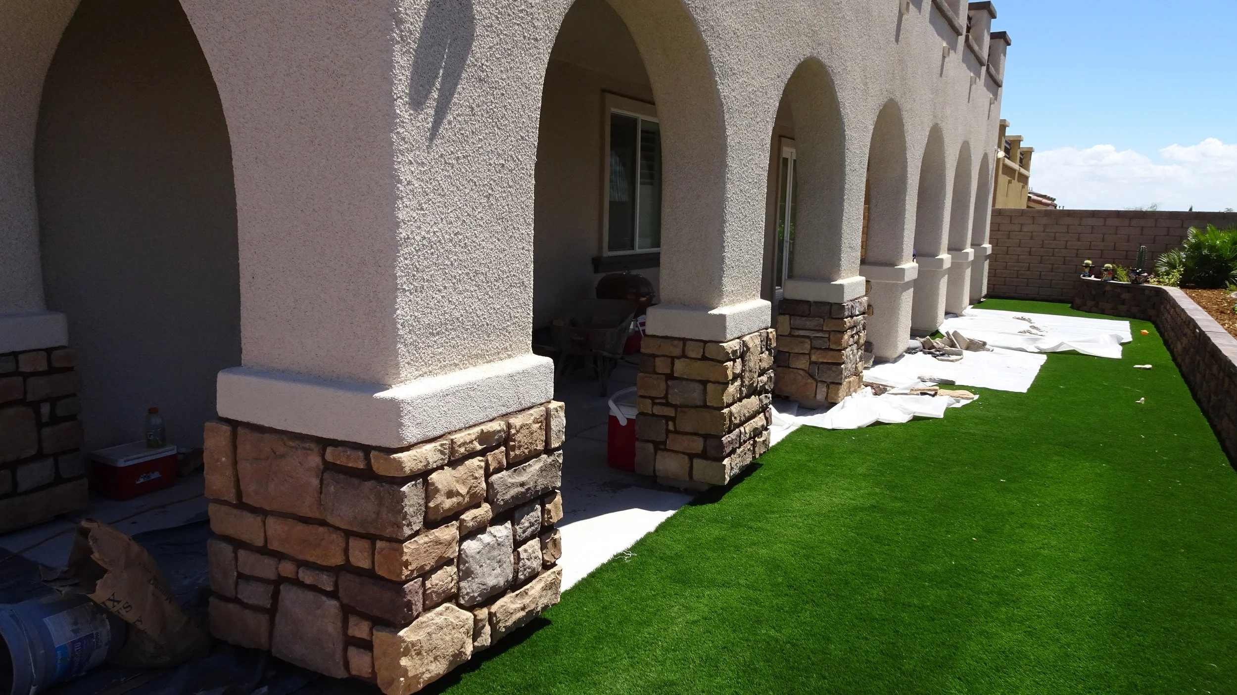 Exterior of a house showing a patio with arches, stone and stucco columns, green artificial grass, and construction supplies under white sheets.