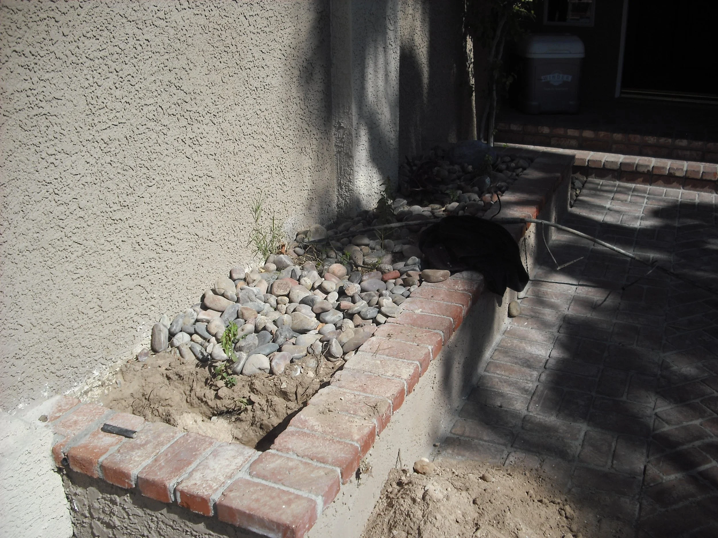 A newly landscaped brick planter box with small stones and a few plants, next to a textured beige wall and adjacent brick-paved walkway.
