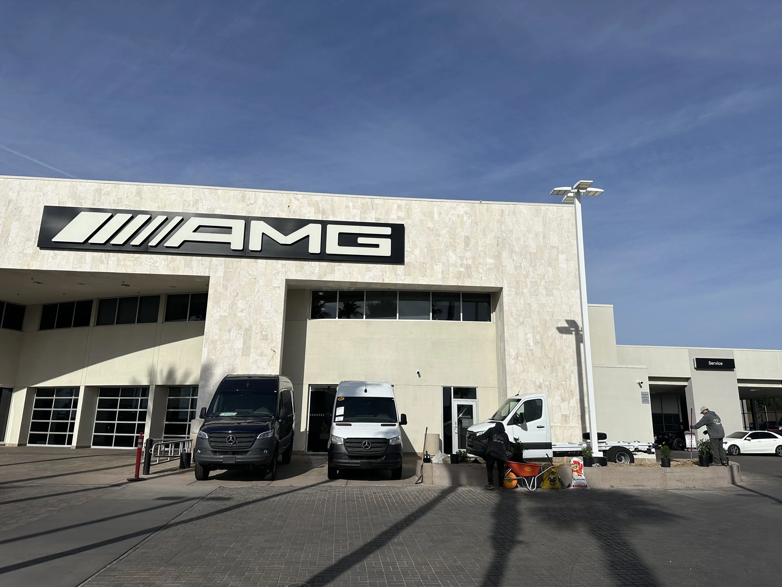 Mercedes-Benz service center with AMG branding, three Mercedes-Benz vans parked in front, and two workers attending to tasks near the sidewalk.