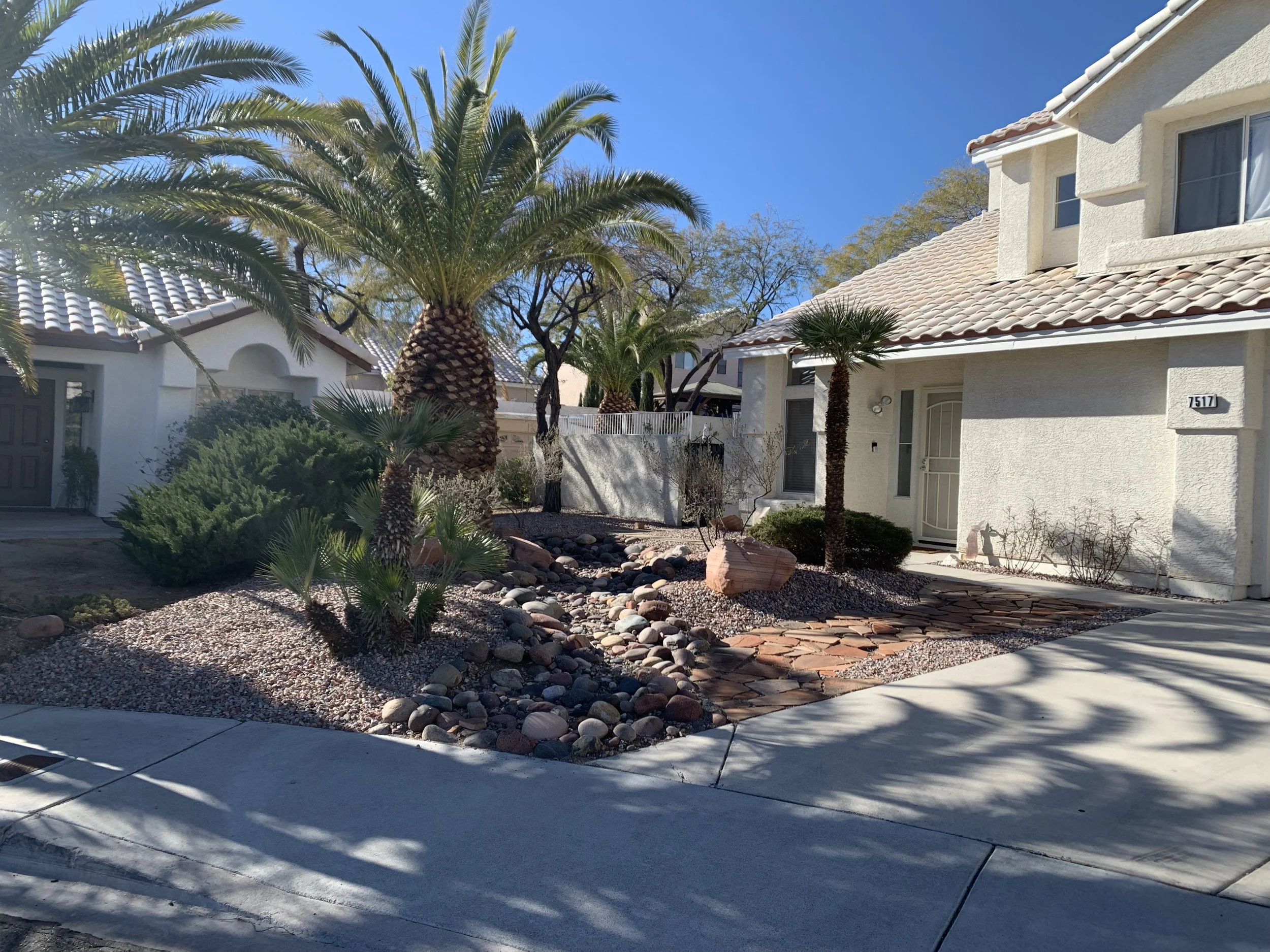 Front yard with desert landscaping, palm trees, rocks, and a white house with a tiled roof in the background.
