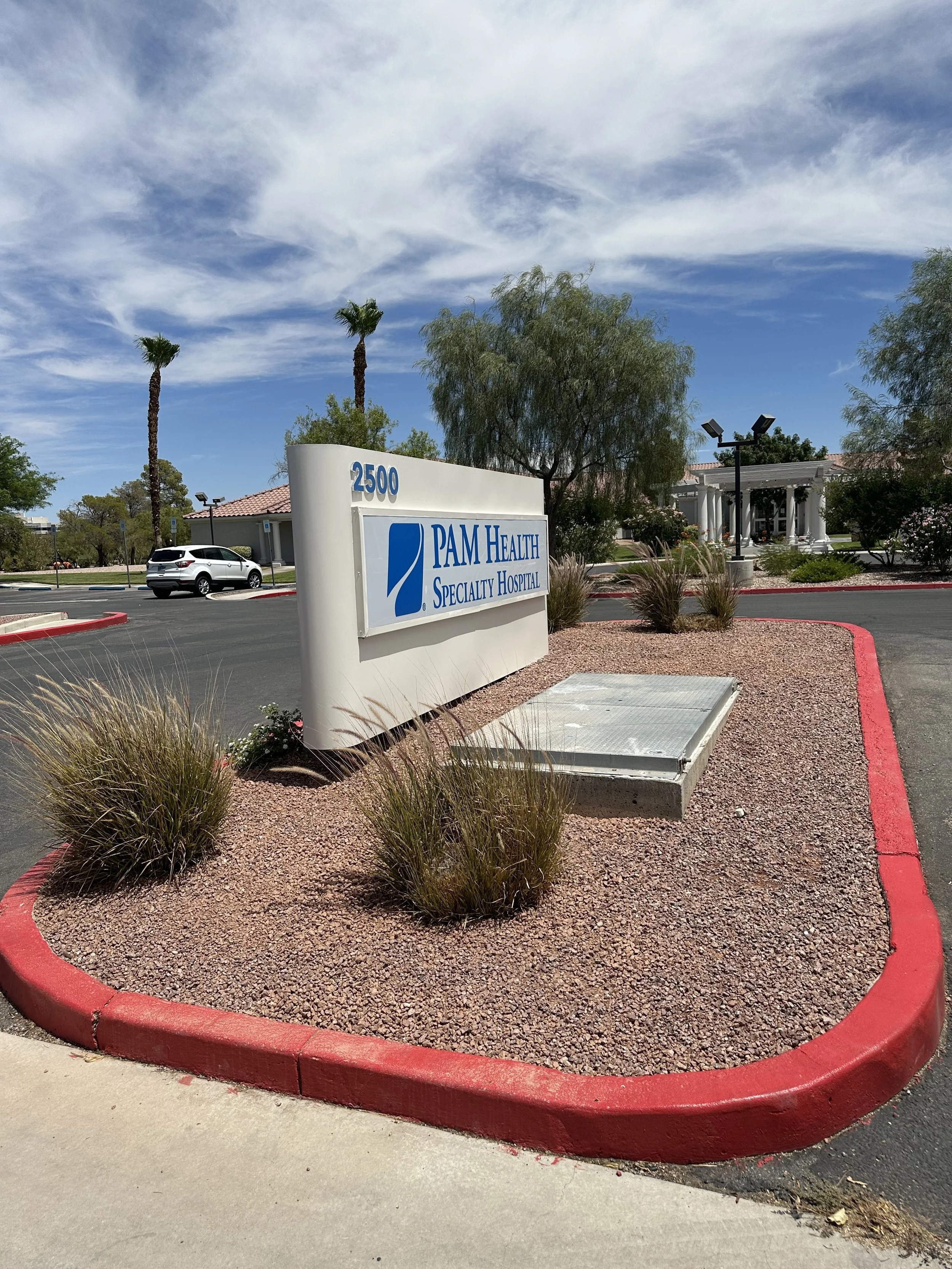 Sign for PAM Health Specialty Hospital at 2500 location, with a landscaped area featuring bushes and a wheelchair ramp, under a partly cloudy sky.