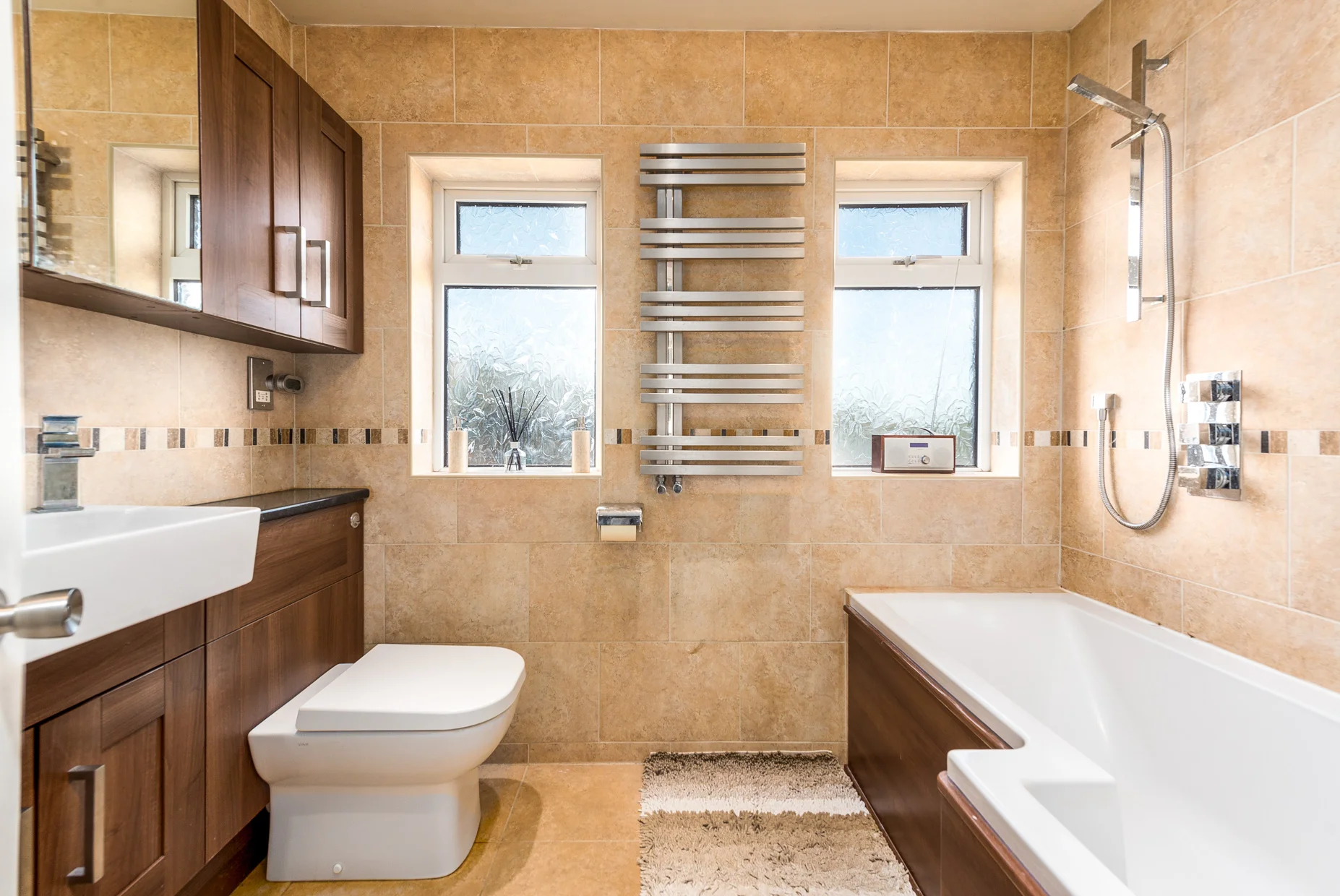 Bathroom with beige tiled walls and floor, two windows, a wooden cabinet with a white sink, a modern toilet, a bath tub with wood paneling, a black countertop, a towel heater, a rug, and various bathroom fixtures.