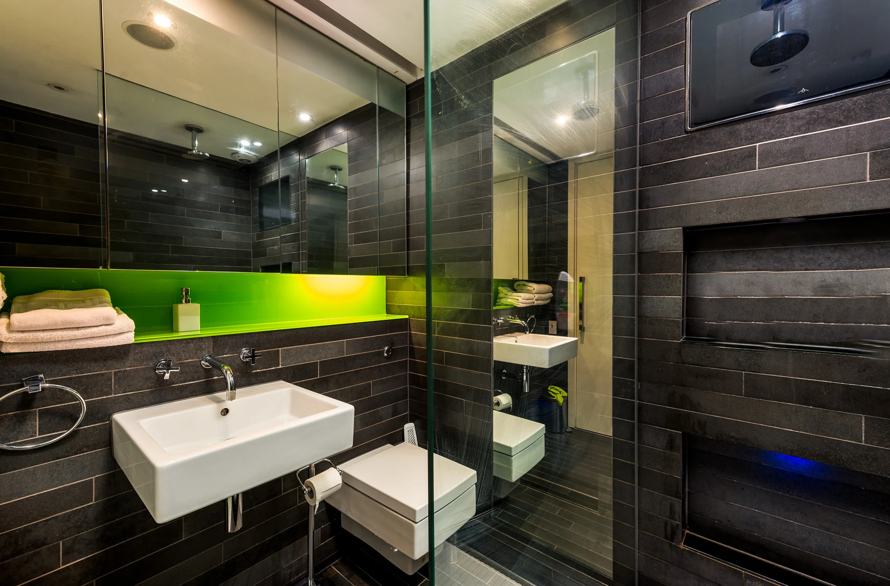 Modern bathroom with black tiled walls, a white sink, a toilet, a small white cabinet, towels, a green illuminated mirror, a glass shower enclosure, and a flat-screen TV mounted on the wall.