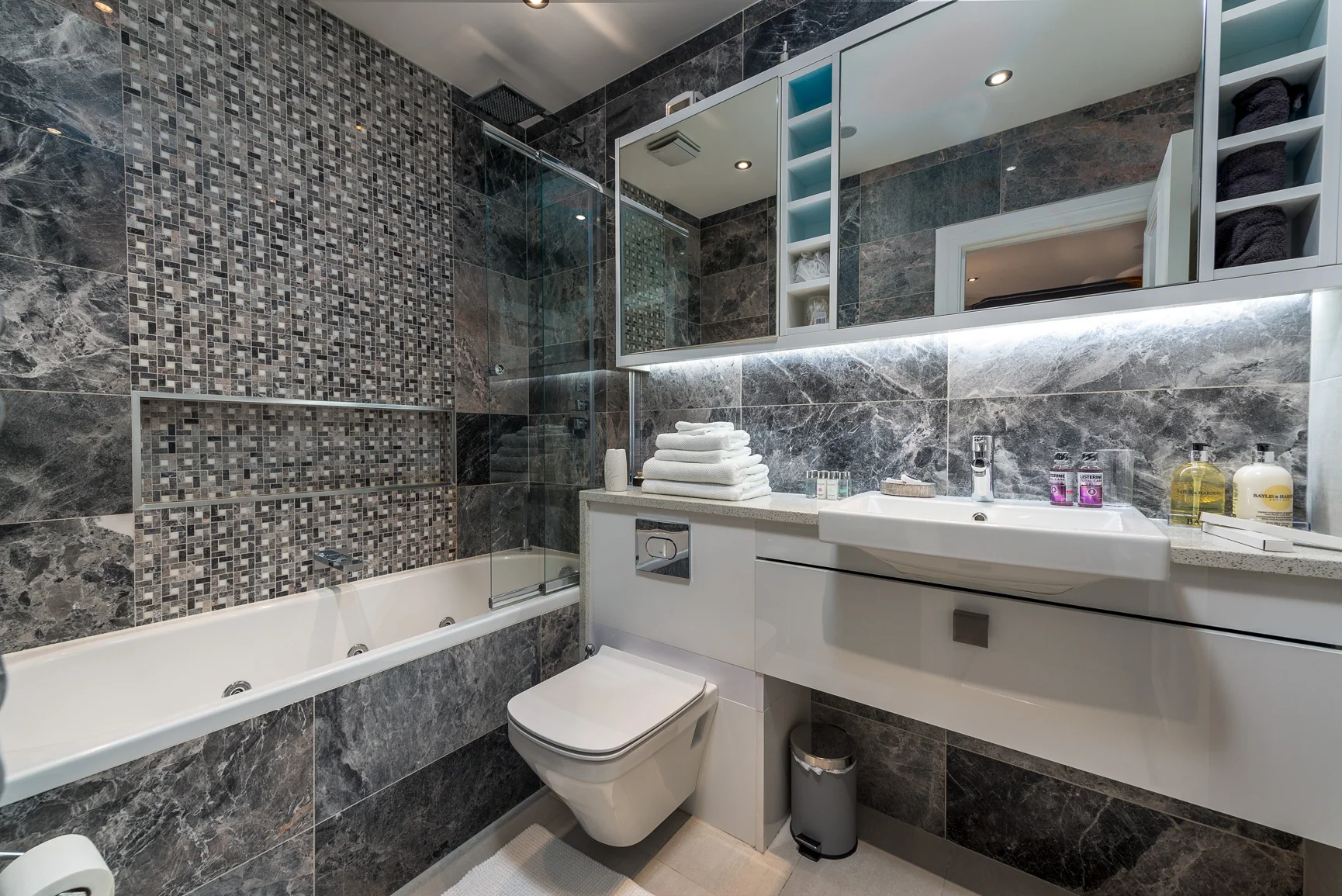 Modern bathroom with dark gray marble tiles, a bathtub with jets, a glass shower door, a white vanity with a sink, mirror cabinets, a stack of white towels, various toiletries, and a small trash bin.