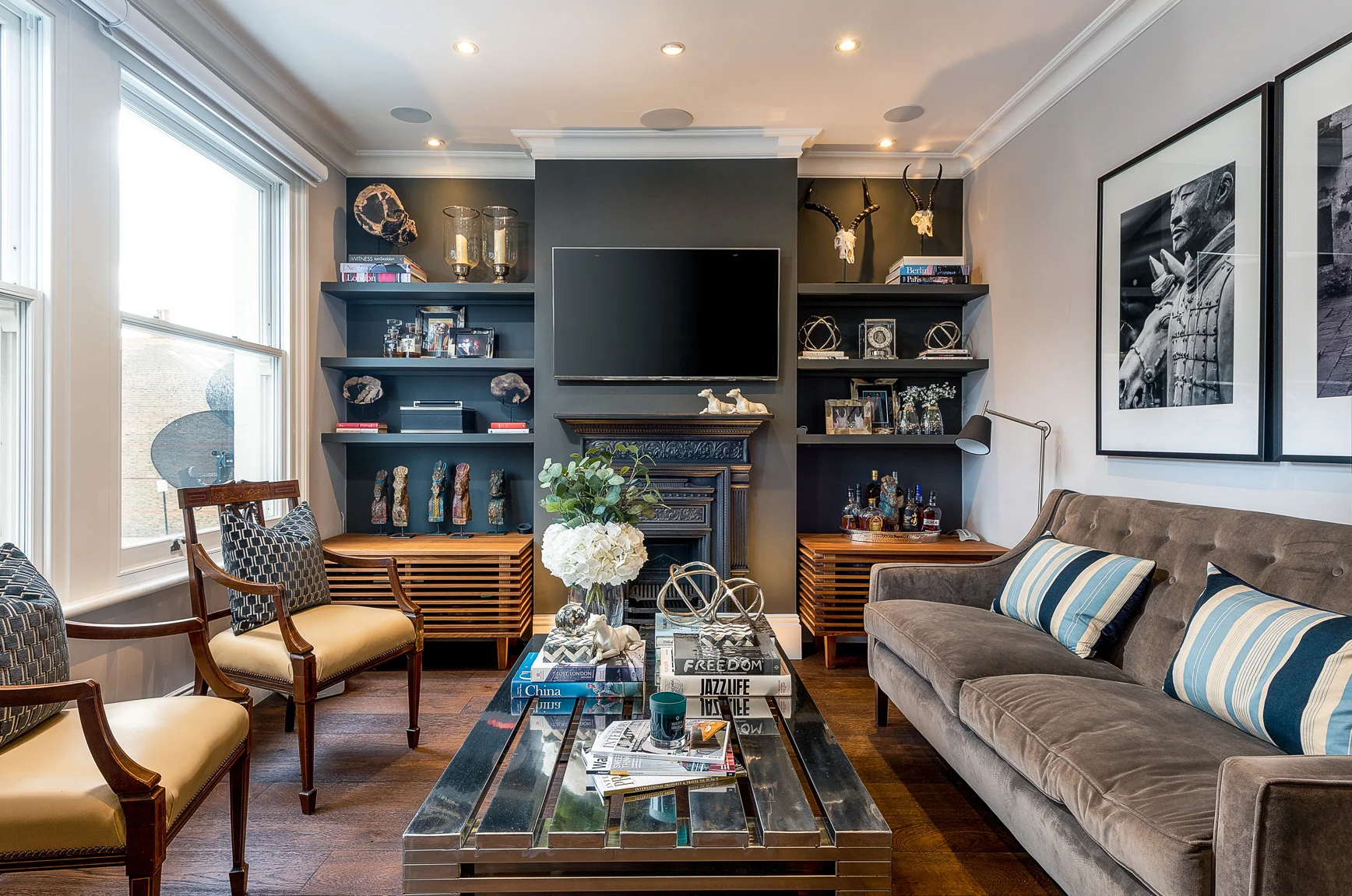 Living room with a gray sofa and armchairs, a black wall with built-in shelves, a flat-screen TV above a decorative fireplace, a glass coffee table with books, and framed black-and-white artwork on the wall.