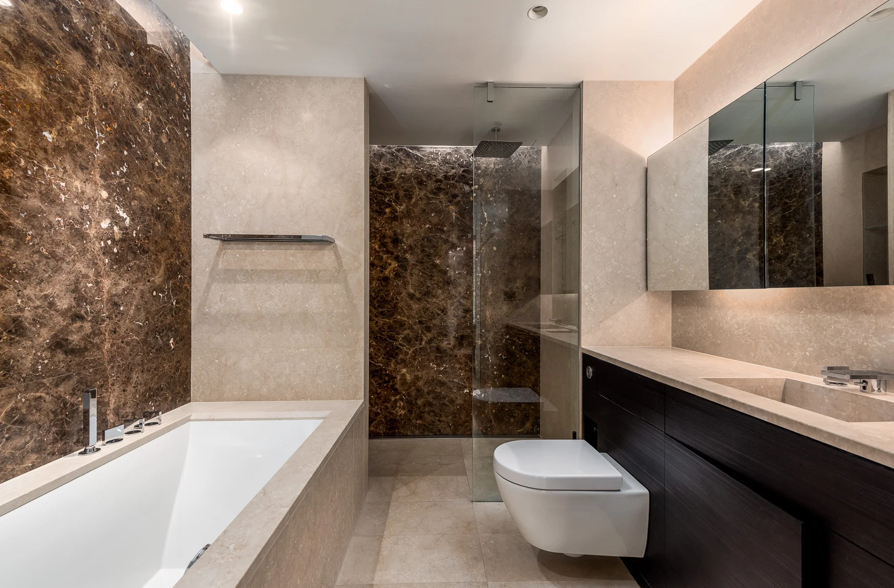 Modern bathroom featuring a built-in bathtub with multiple faucet controls, a glass-enclosed shower area with a rain showerhead, a toilet, a large mirror, and beige and dark brown marble and stone finishes.