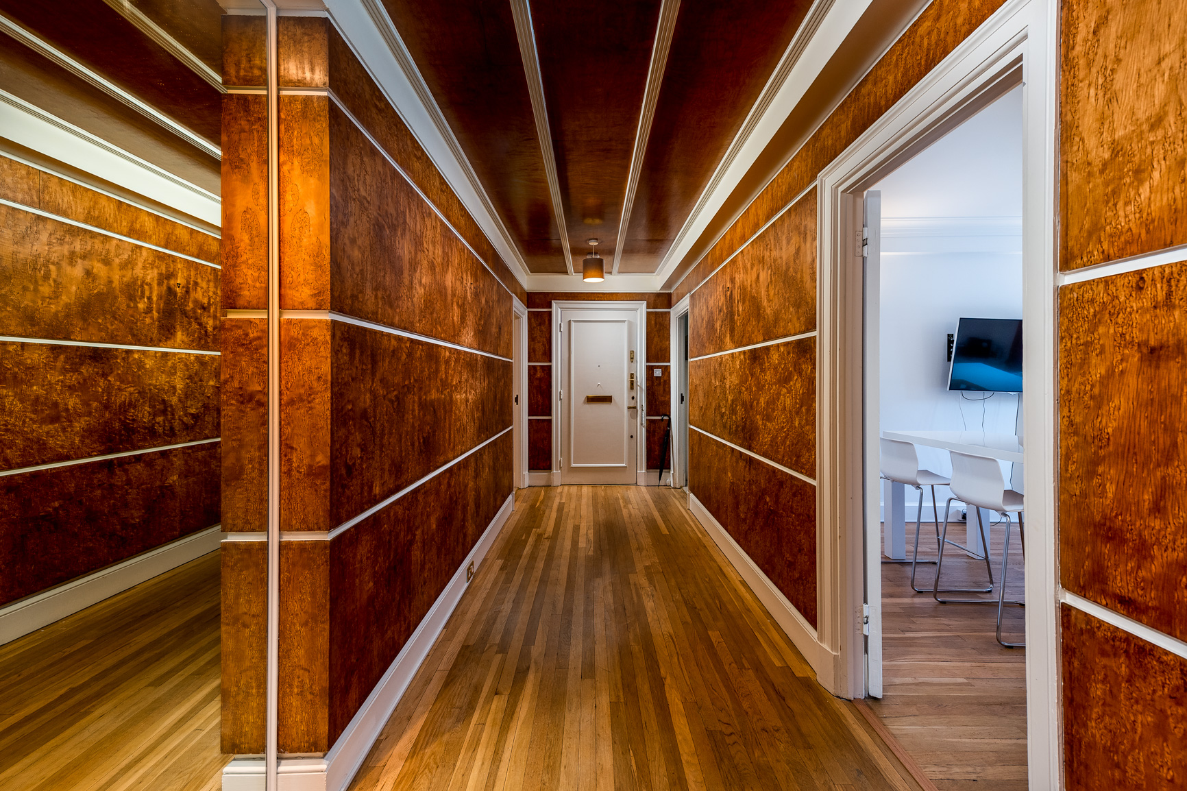 A hallway with wooden floors and walls adorned with wood paneling, leading to a white front door. To the right, an open doorway reveals a room with white walls, a mounted TV, and a white table with white chairs.
