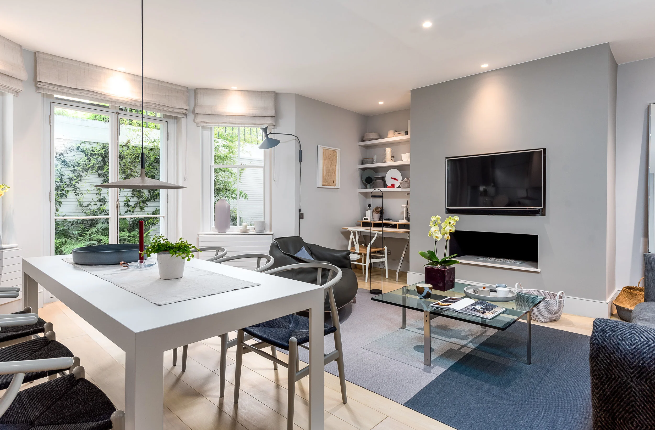 Modern living room with large windows, white dining table, black sofa, flat-screen TV on light gray wall, glass coffee table, and open shelves with decorative items.