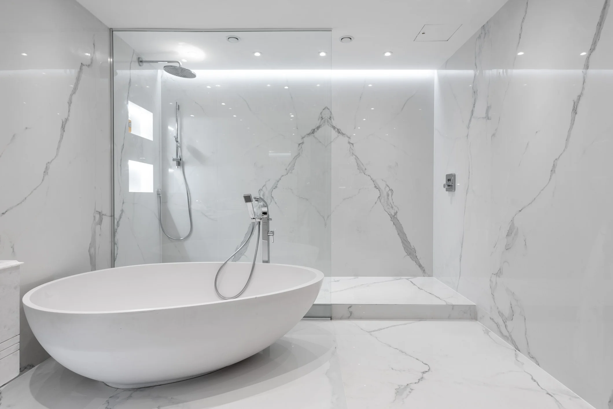 Modern minimalist bathroom with white marble walls and floor, featuring a freestanding oval bathtub, a glass shower enclosure with a rainfall showerhead and handheld shower, and sleek fixtures.