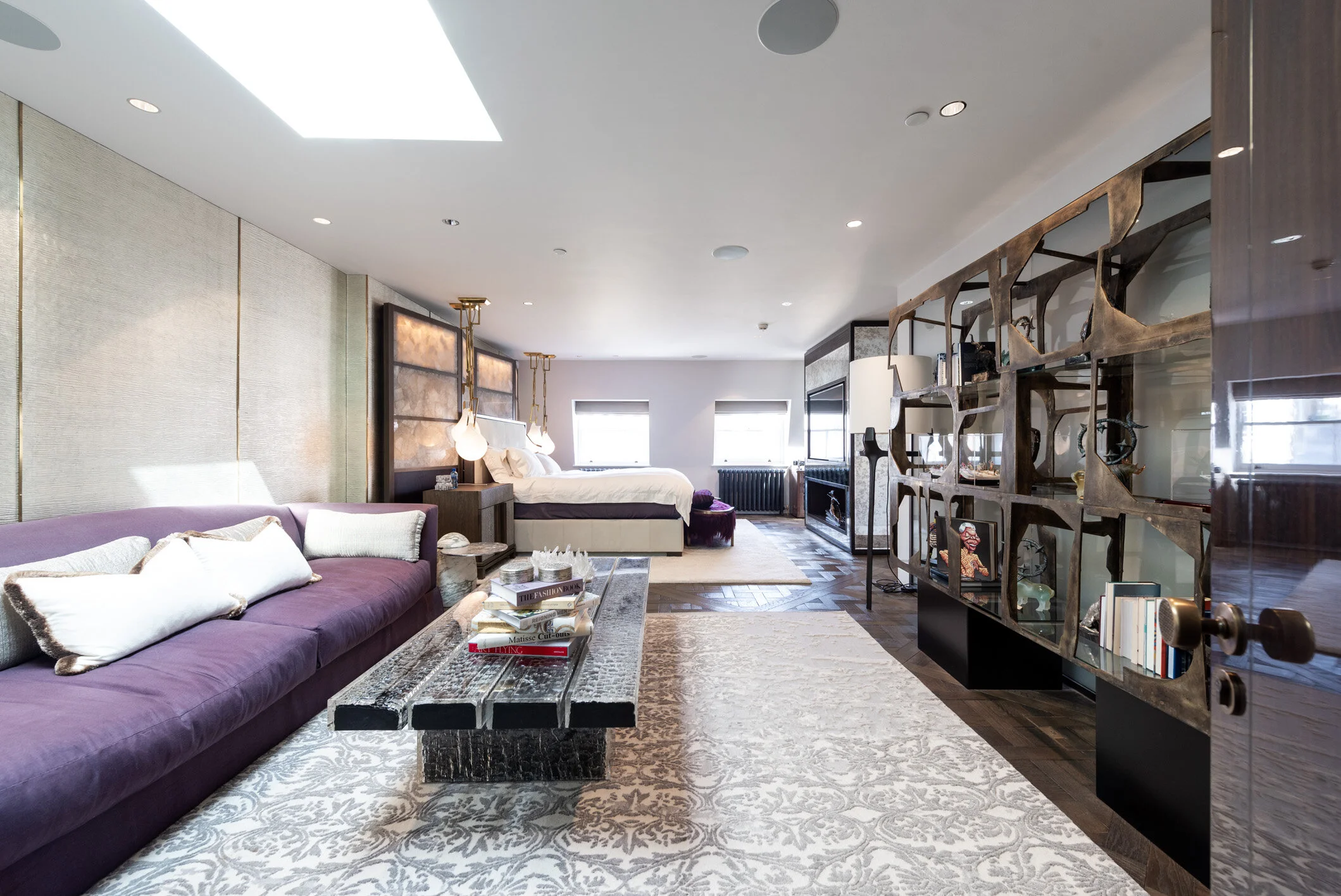Modern living room with purple sofa, coffee table, area rug, and a bedroom in the background with a bed and windows, separated by an open decorative shelving unit.