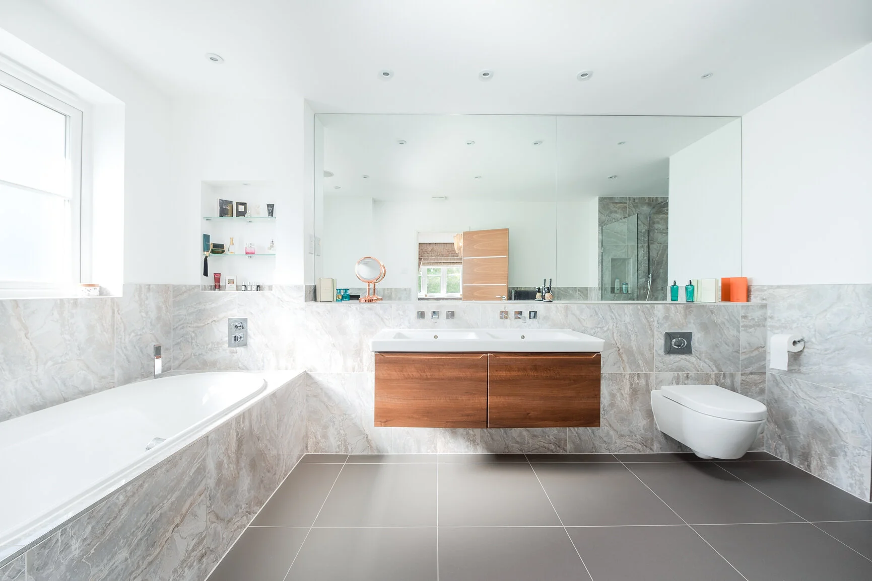 Modern white bathroom with a bathtub, a wooden vanity with a sink, a large mirror, a wall-mounted toilet, and a shower in the background, with natural light coming through the window.