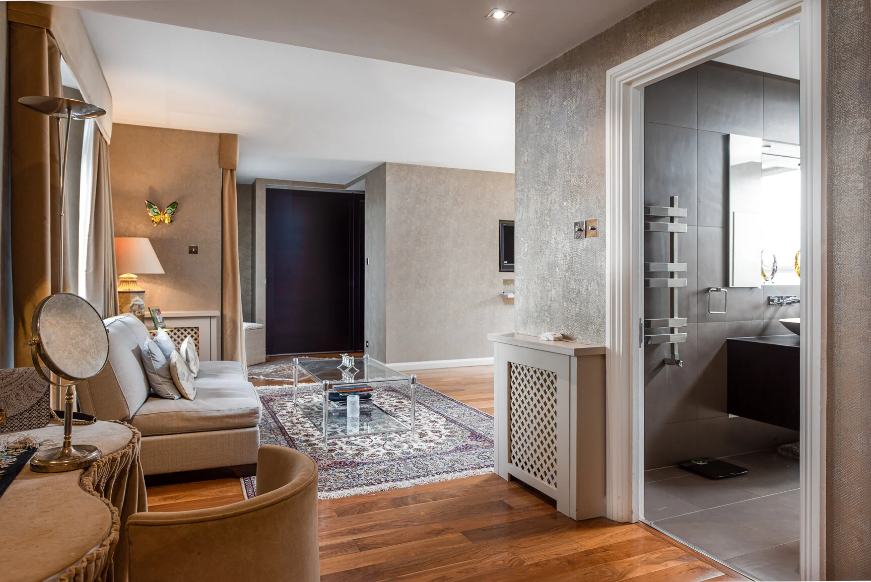 Living room with beige sofa, glass coffee table, area rug, and wall-mounted TV; doorway to bathroom with tiled walls and modern fixtures; warm lighting and decorative elements.