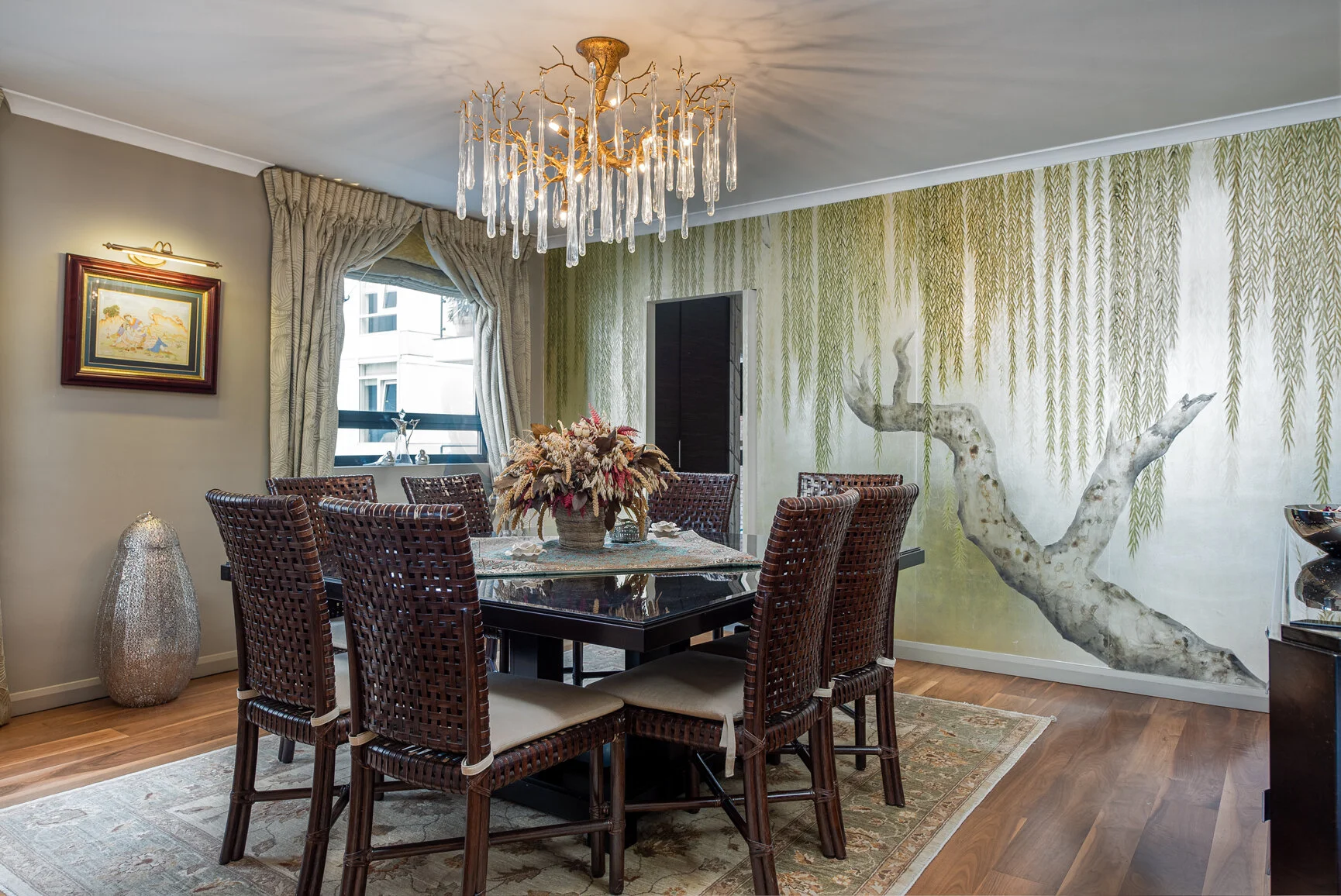 Dining room with a dark wooden table, six woven wooden chairs with beige cushions, and a floral centerpiece. There is a large window with patterned curtains, a crystal chandelier hanging from the ceiling, a decorative vase on the floor, and a wall mu