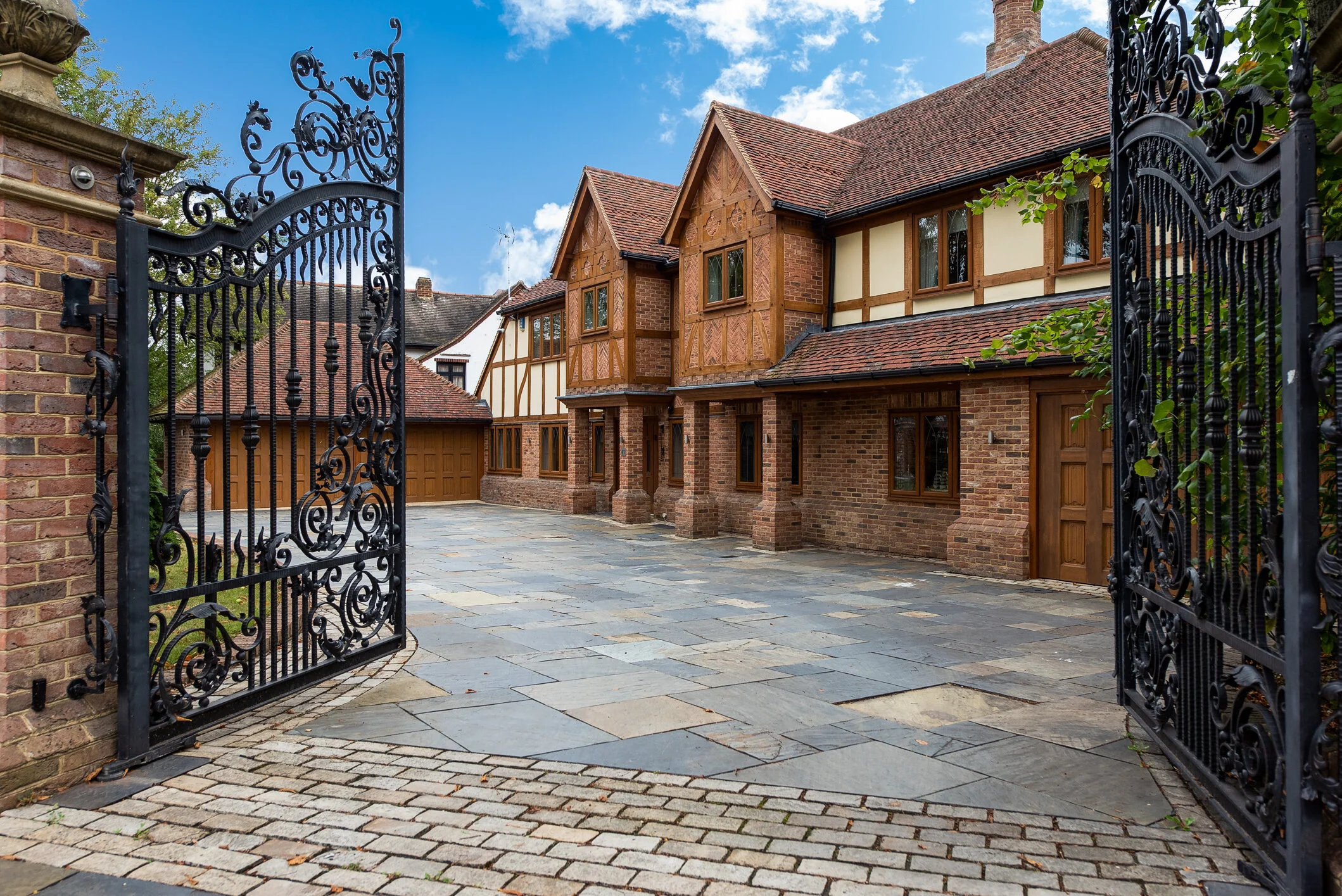 A large, elegant brick and wood house viewed through black wrought iron gates, with a cobblestone driveway and a partly cloudy sky.
