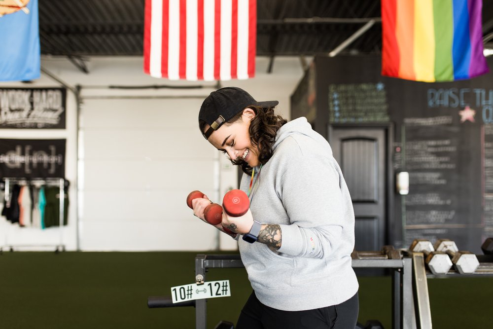 okc oklahoma norman commercial photography woman with backwards hat and brown hair with tattoos lifting weights in the gym while laughing headshots branding photography