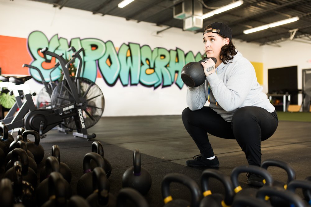 okc oklahoma norman commercial photography woman with backwards hat and brown hair with tattoos doing squats in the gym with kettlebell headshots branding photography