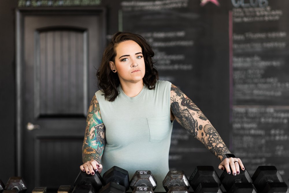 okc oklahoma norman commercial photography woman with backwards hat and brown hair with tattoos standing in front of weights in the gym looking at the camera headshots branding photography