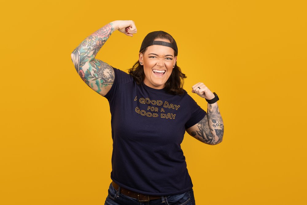 okc oklahoma norman commercial photography woman with backwards hat and brown hair with tattoos on yellow background making strong arm pose  headshots branding photography
