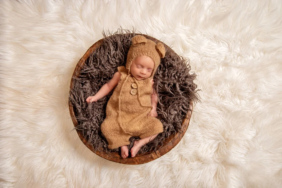 A newborn baby sleeping in a round wooden bowl on a fluffy white rug, wearing a brown knit bear costume with bear ears on the hat.