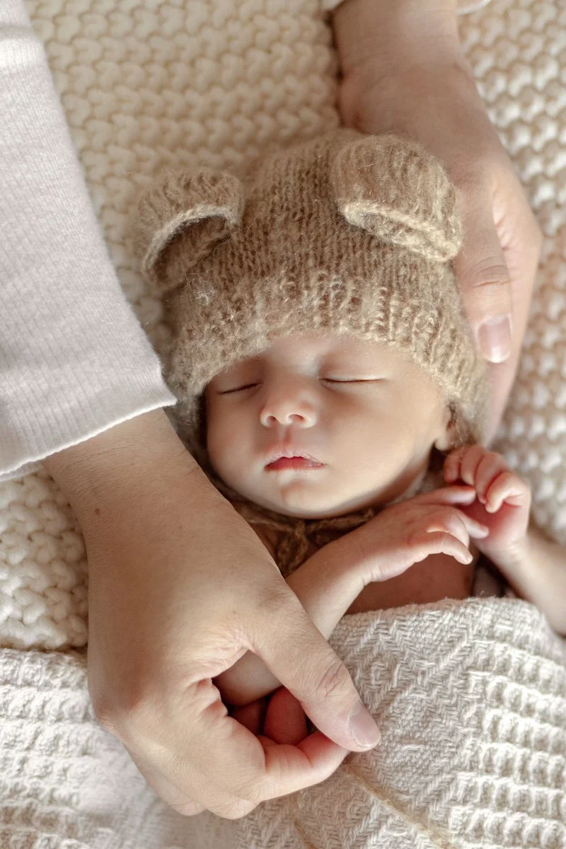 Close-up of a newborn baby peacefully sleeping, wearing a cozy bear-themed knit hat, cradled gently in adult hands on a soft textured blanket.