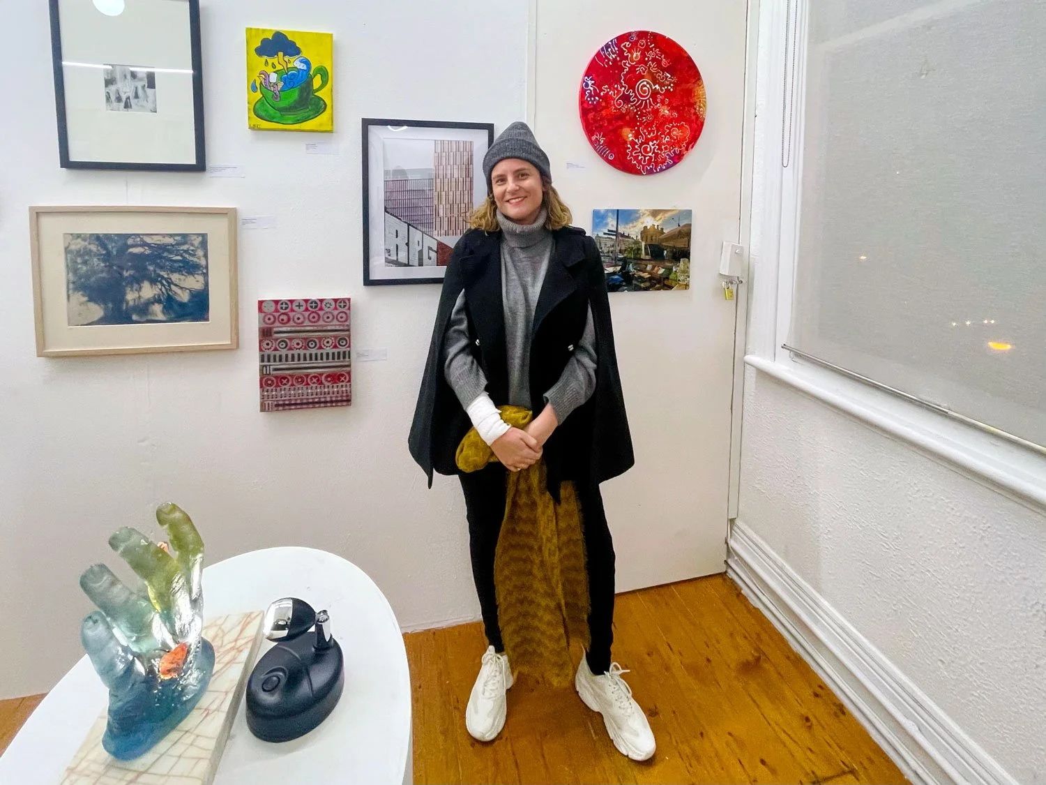 Person in a grey beanie and black cape stands against a white gallery wall hung with mixed artworks; in the foreground, a white pedestal holds a blue‑green glass hand sculpture and a small black object.