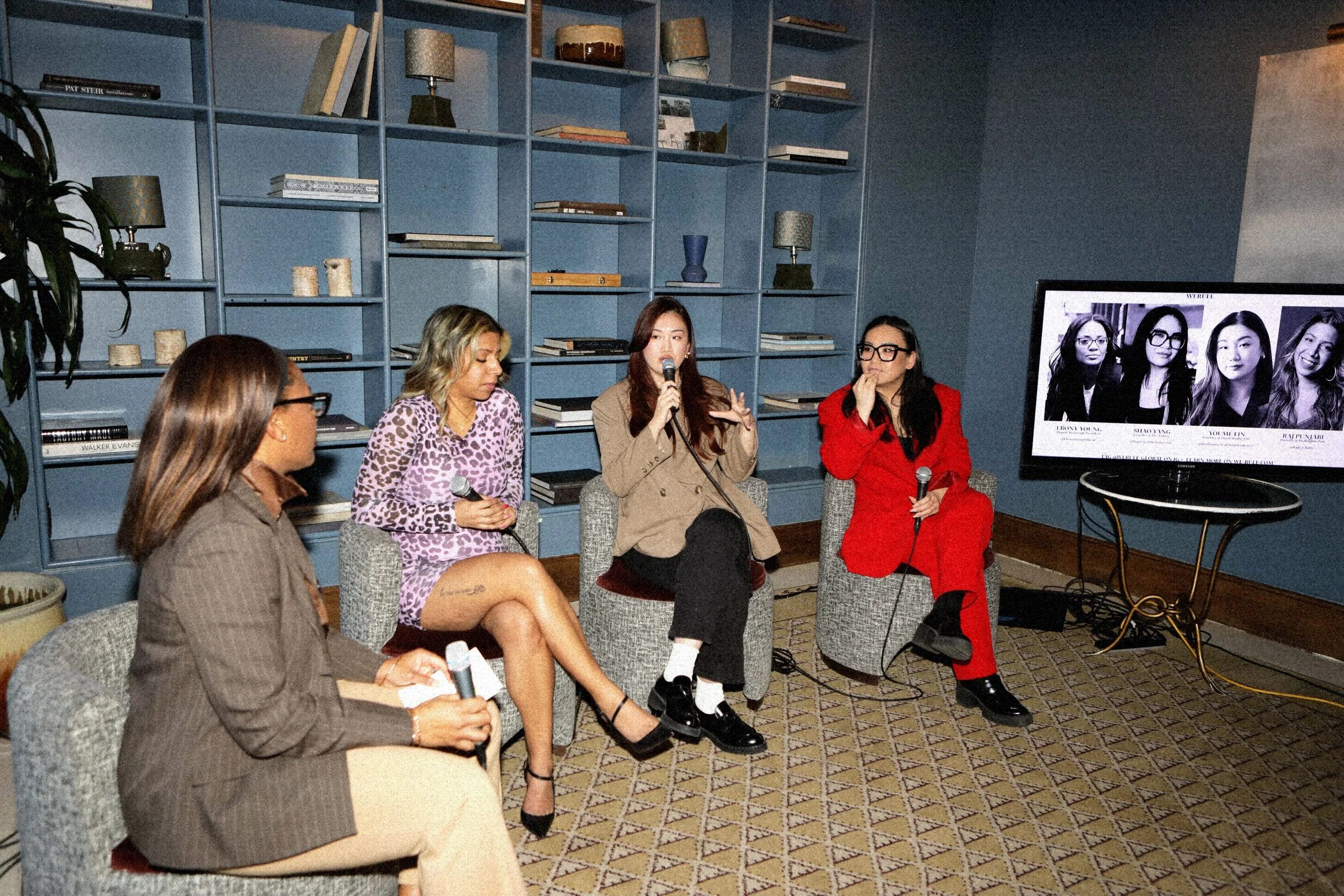Five women sitting on chairs in a panel discussion, with a bookshelf behind them and a monitor displaying headshots of the panelists.