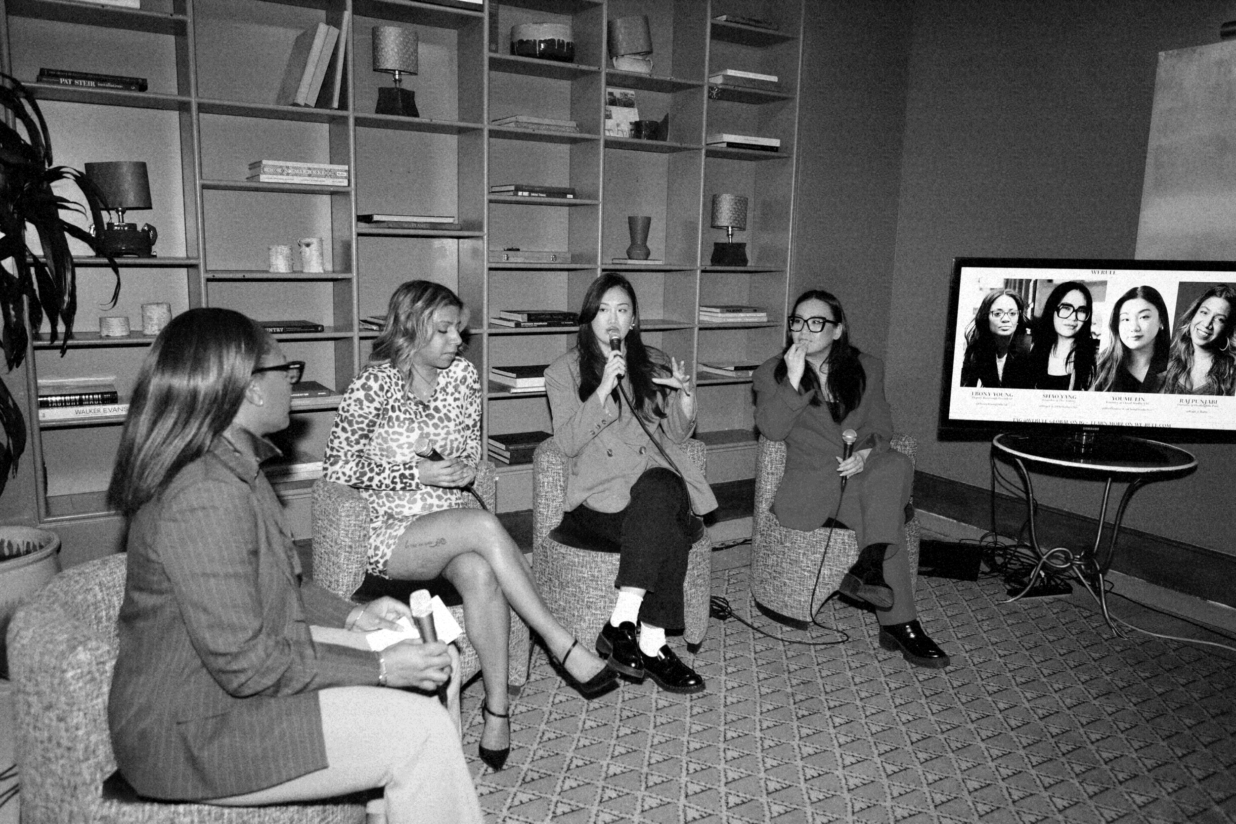 Four women seated on chairs participating in a panel discussion in a room with shelves filled with books and decorative items. One woman is speaking into a microphone, while the others listen. A television screen on the right displays headshots of additional women.