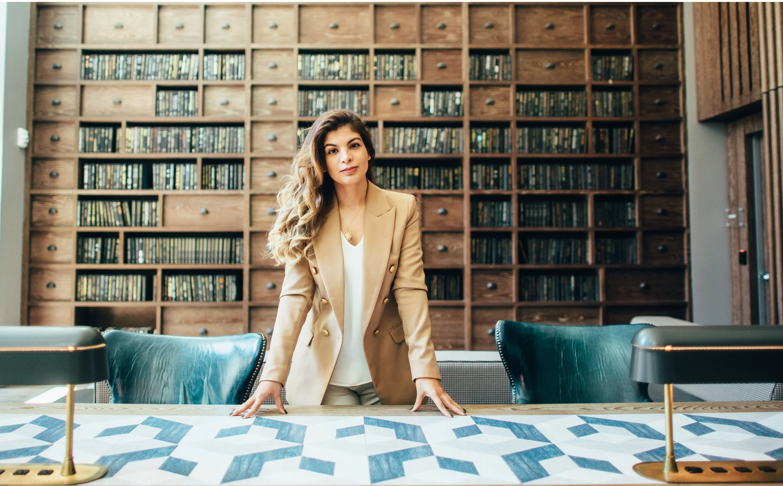 Woman in beige blazer standing at a conference table in a library or office with wooden bookshelves and leather chairs.