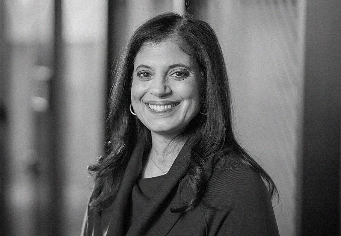 Black and white portrait of a smiling woman with long dark hair, wearing earrings and a dark top, standing against a blurred background.