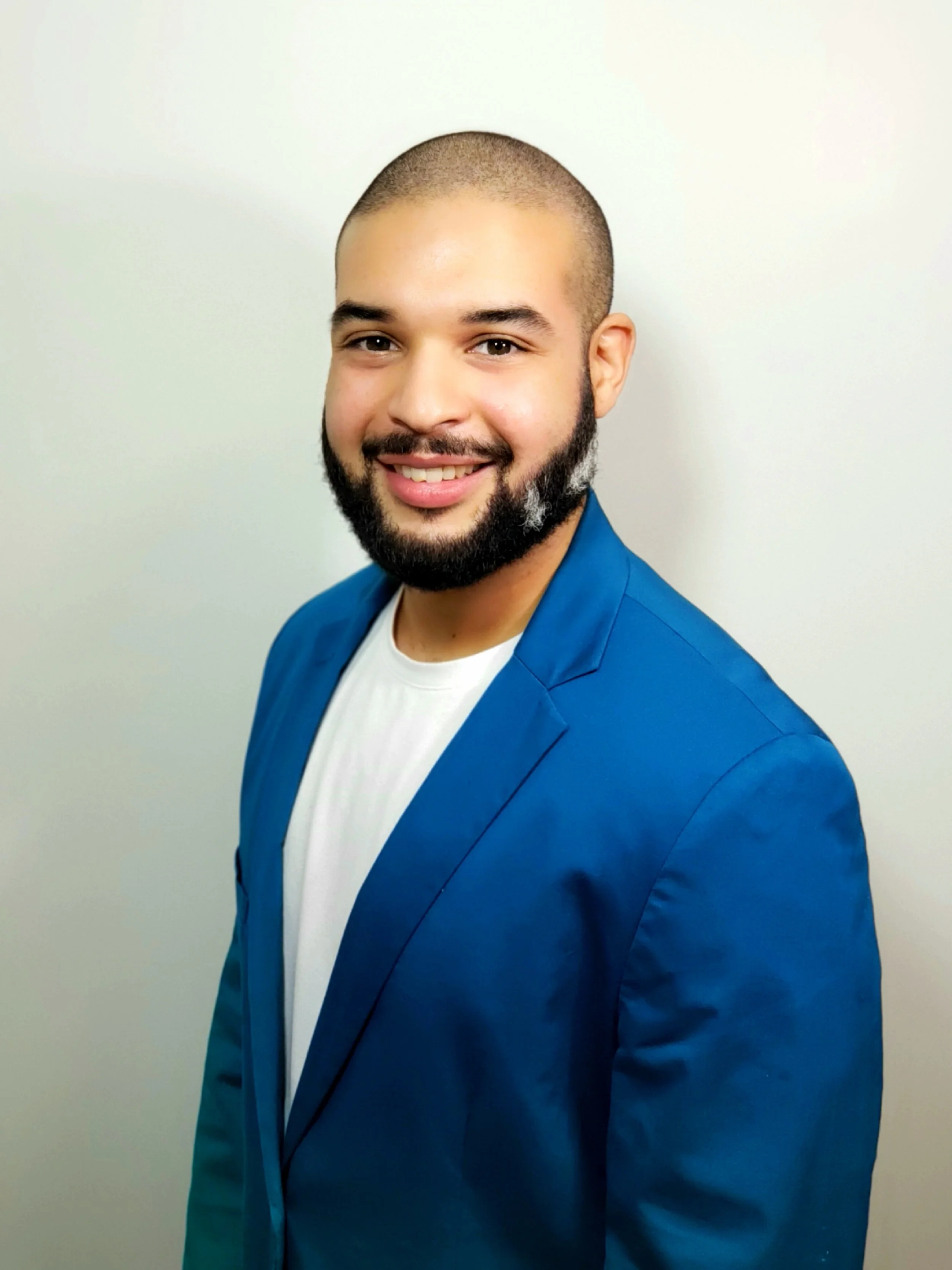 A young man with a beard, smiling, wearing a blue blazer over a white shirt, standing against a plain white background.