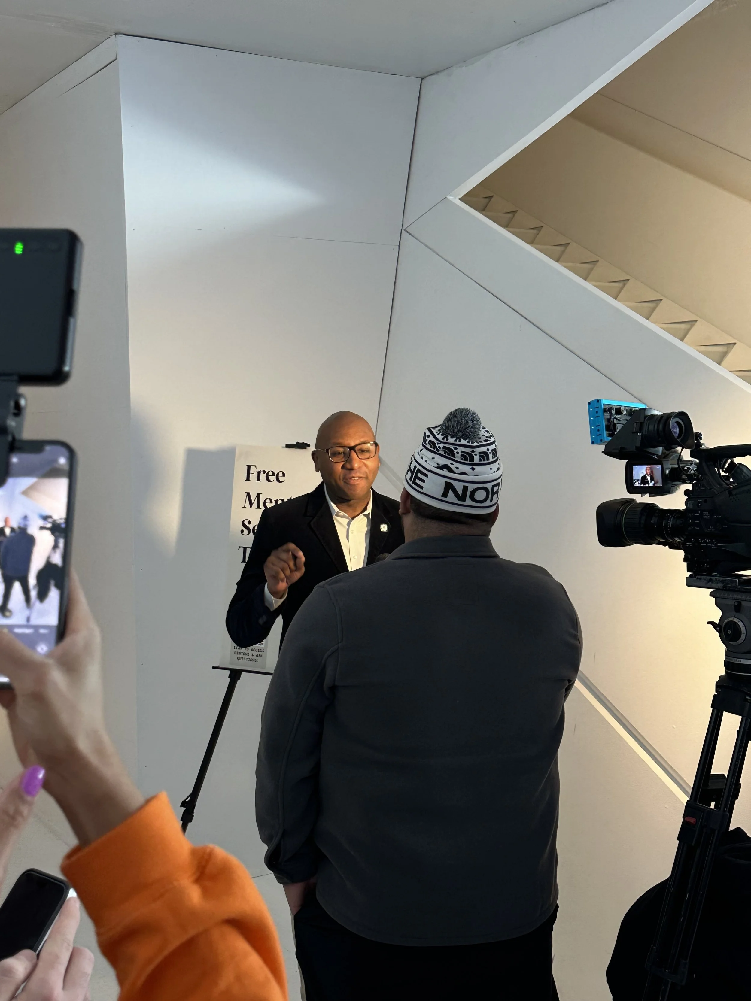 A man in a suit and glasses speaking to a person wearing a beanie hat at a media interview or press event indoors with a sign in the background that reads 'Free Ment...'