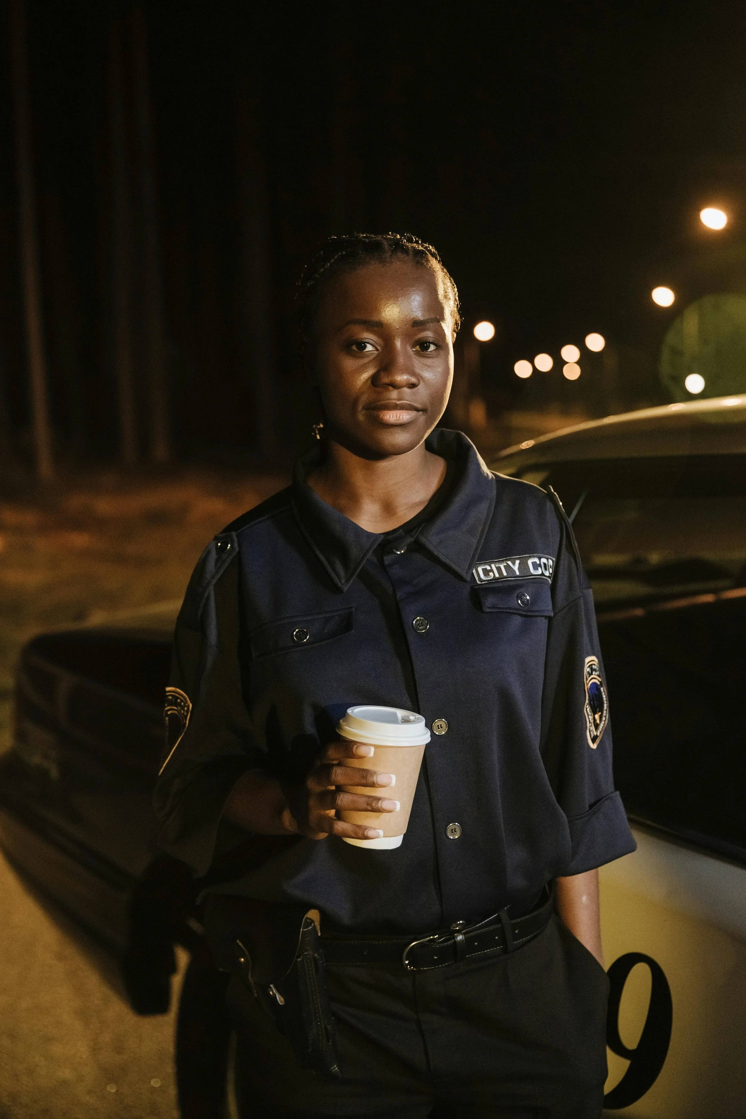 Nighttime photo of a female police officer in uniform holding a disposable coffee cup, standing beside a police vehicle.