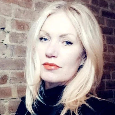 A woman with blonde hair, wearing a black top, standing in front of a brick wall.