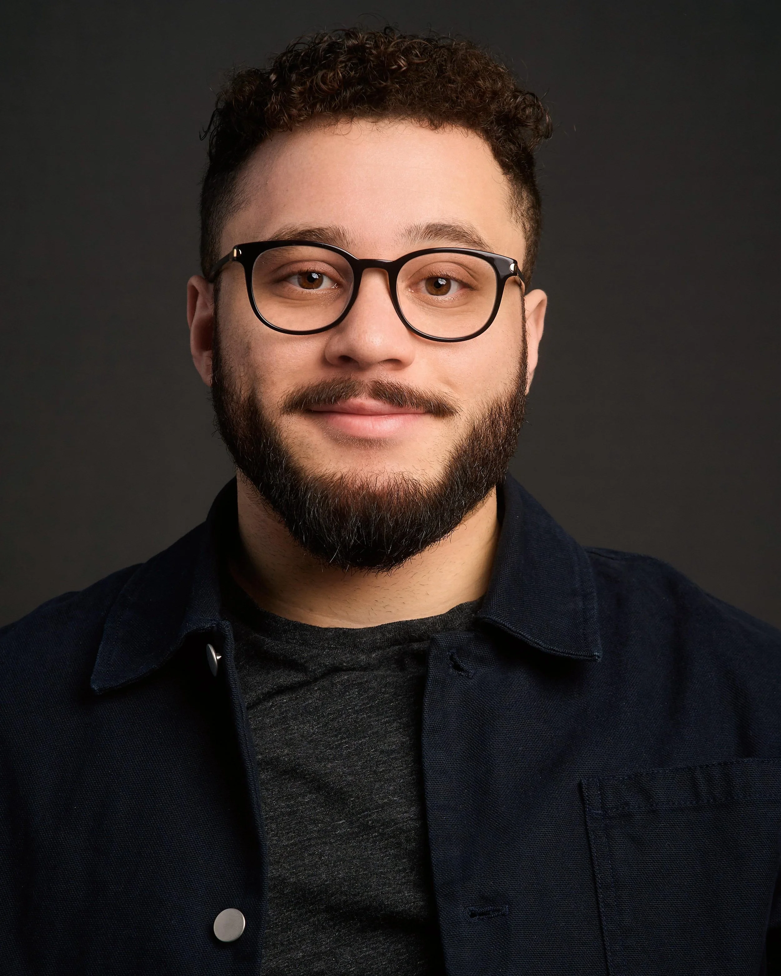 A young man with curly hair, glasses, and a beard smiling, wearing a dark jacket over a gray shirt against a dark background.