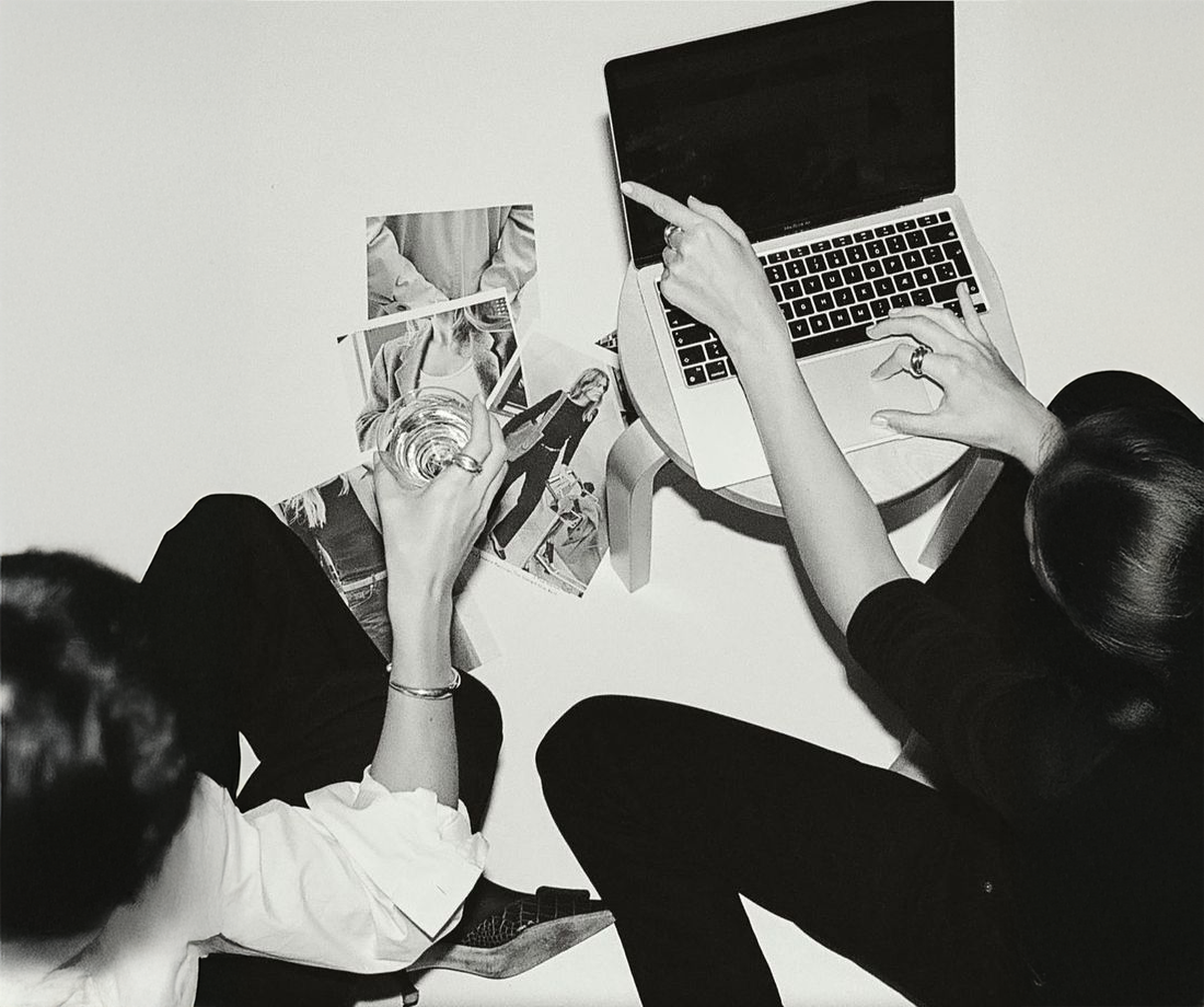 Two women sitting at a white table, one using a laptop and the other looking at photos, with a glass of water on the table.