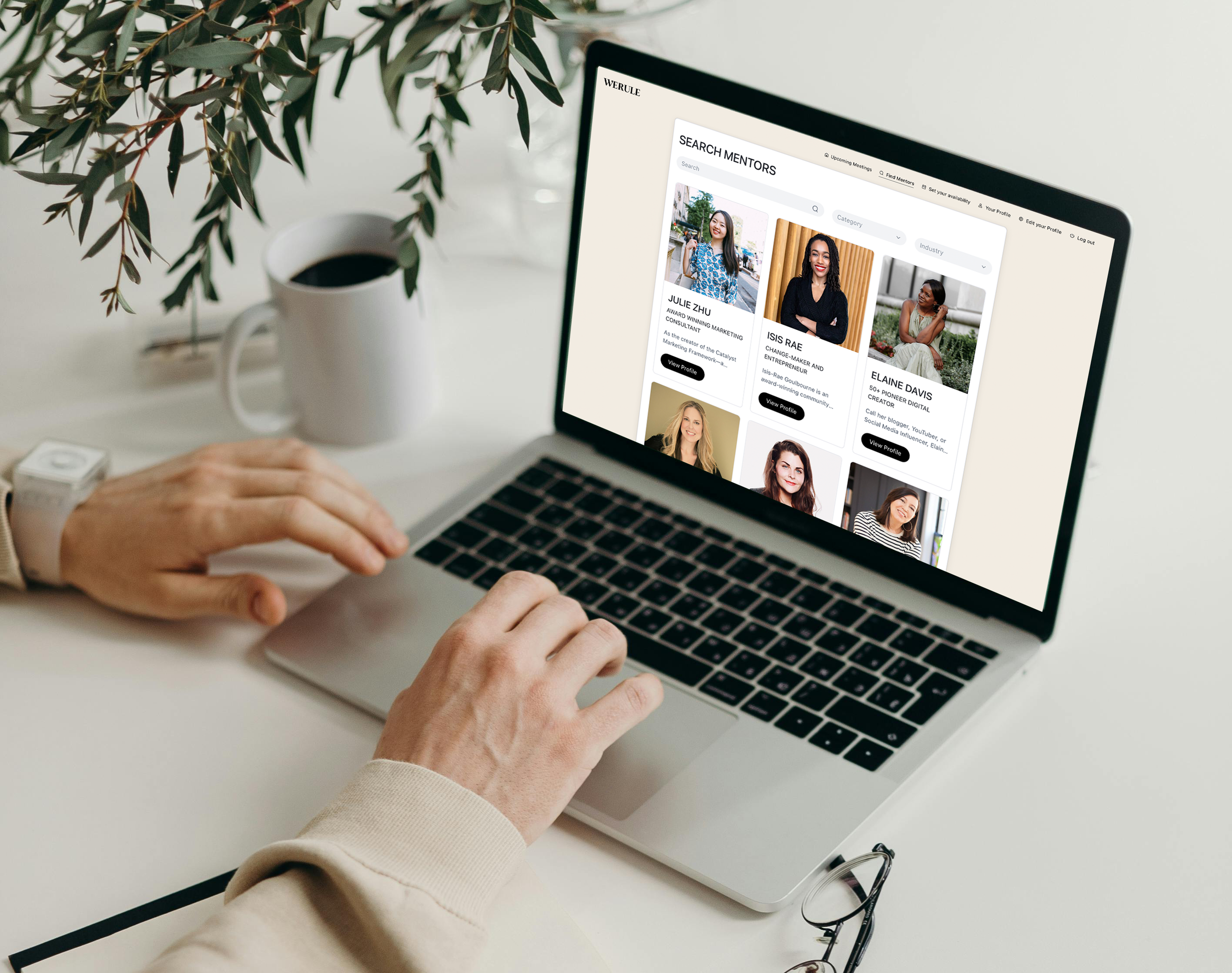 Person using a laptop on a desk displaying a mentor search website, with profiles and photos of mentors. A cup of coffee, eyeglasses, and a notebook are on the desk. Leaves are in the foreground.