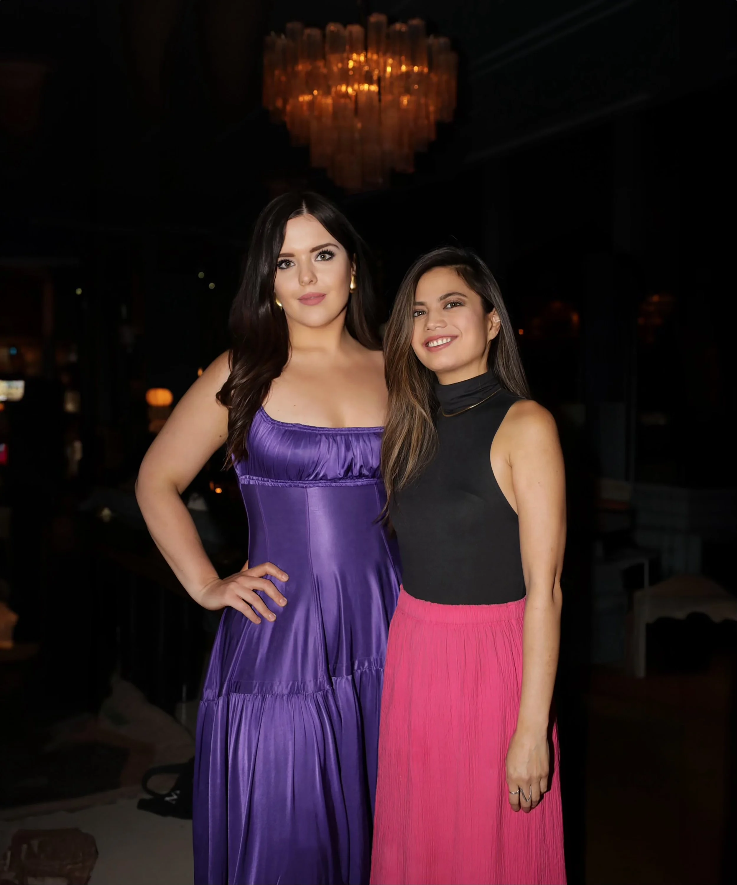 Two women posing together indoors, one wearing a purple satin dress and the other wearing a black sleeveless top with a pink skirt. They are smiling and standing close to each other with a chandelier above them.